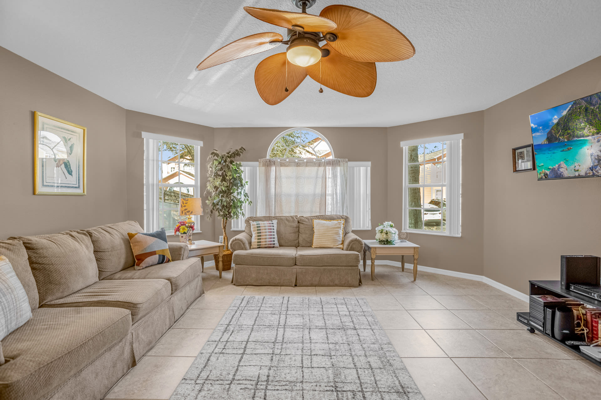 A wide shot of the living room, showcasing two beige sofas, a patterned rug, a large ceiling fan, and multiple windows with arched tops, creating a bright and airy space.