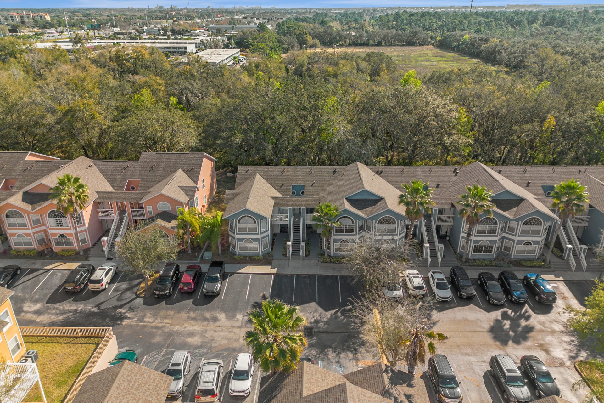 An aerial perspective of a residential complex, showcasing rows of light blue and orange multi-story buildings with pitched roofs, interspersed with mature trees and extensive parking areas, under a clear sky.