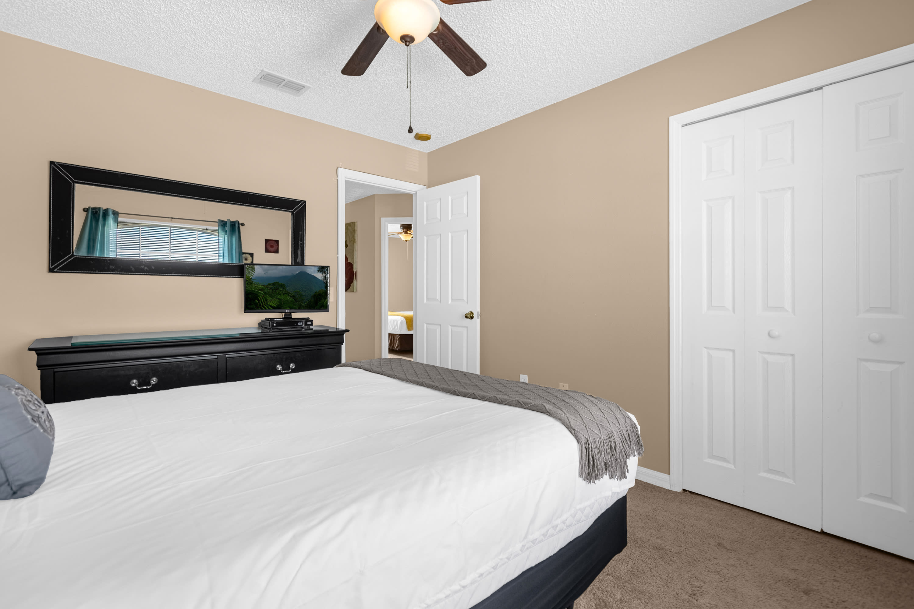 A cozy bedroom features a queen-sized bed with white bedding and a gray throw, a black dresser with a TV, and a large mirror reflecting teal blinds. The room has light beige walls and a ceiling fan.