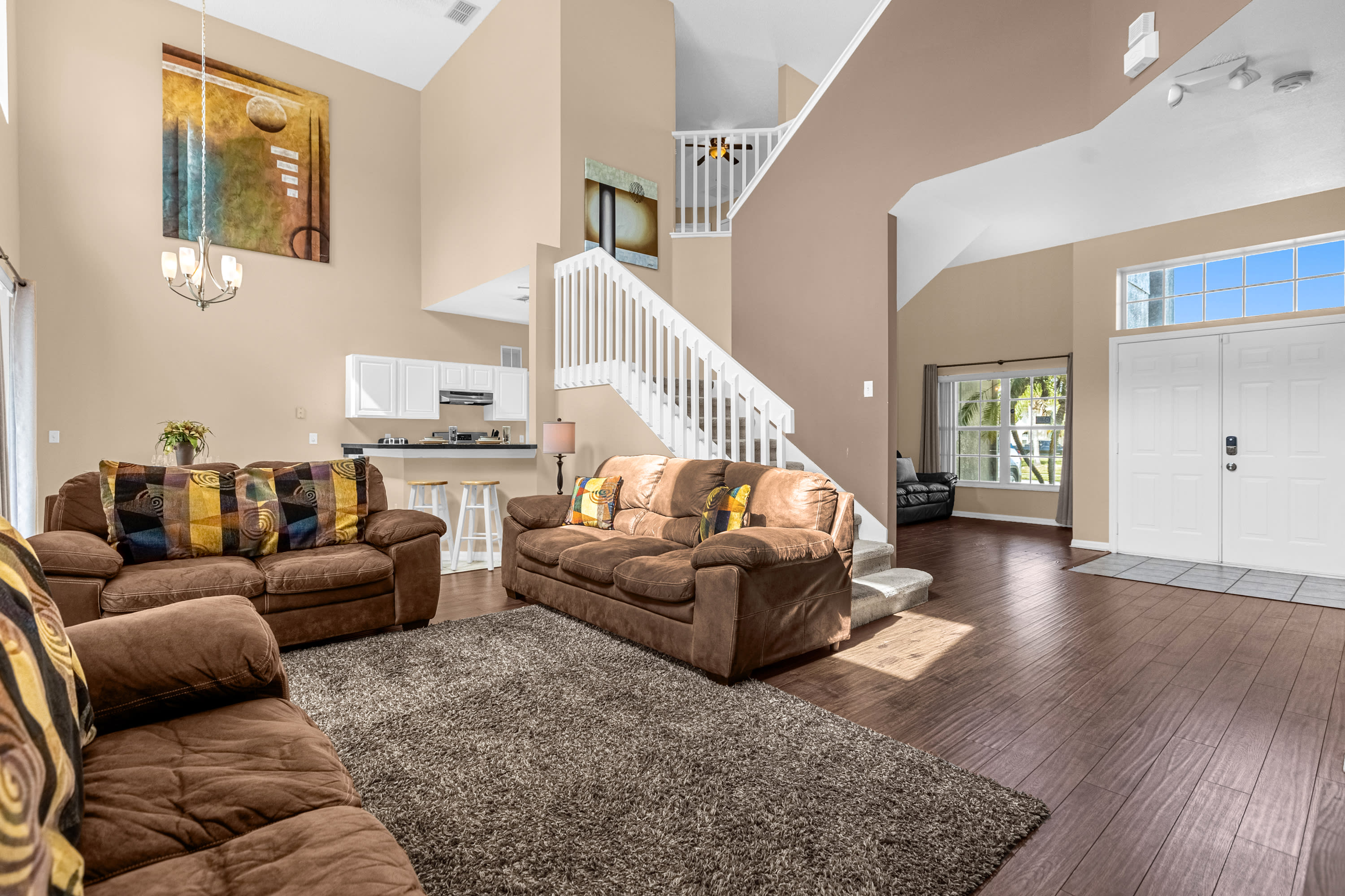 Spacious open-concept living room with three brown leather sofas, a shaggy brown area rug, and dark wood laminate flooring, featuring a high ceiling and a view of the kitchen and a white staircase.