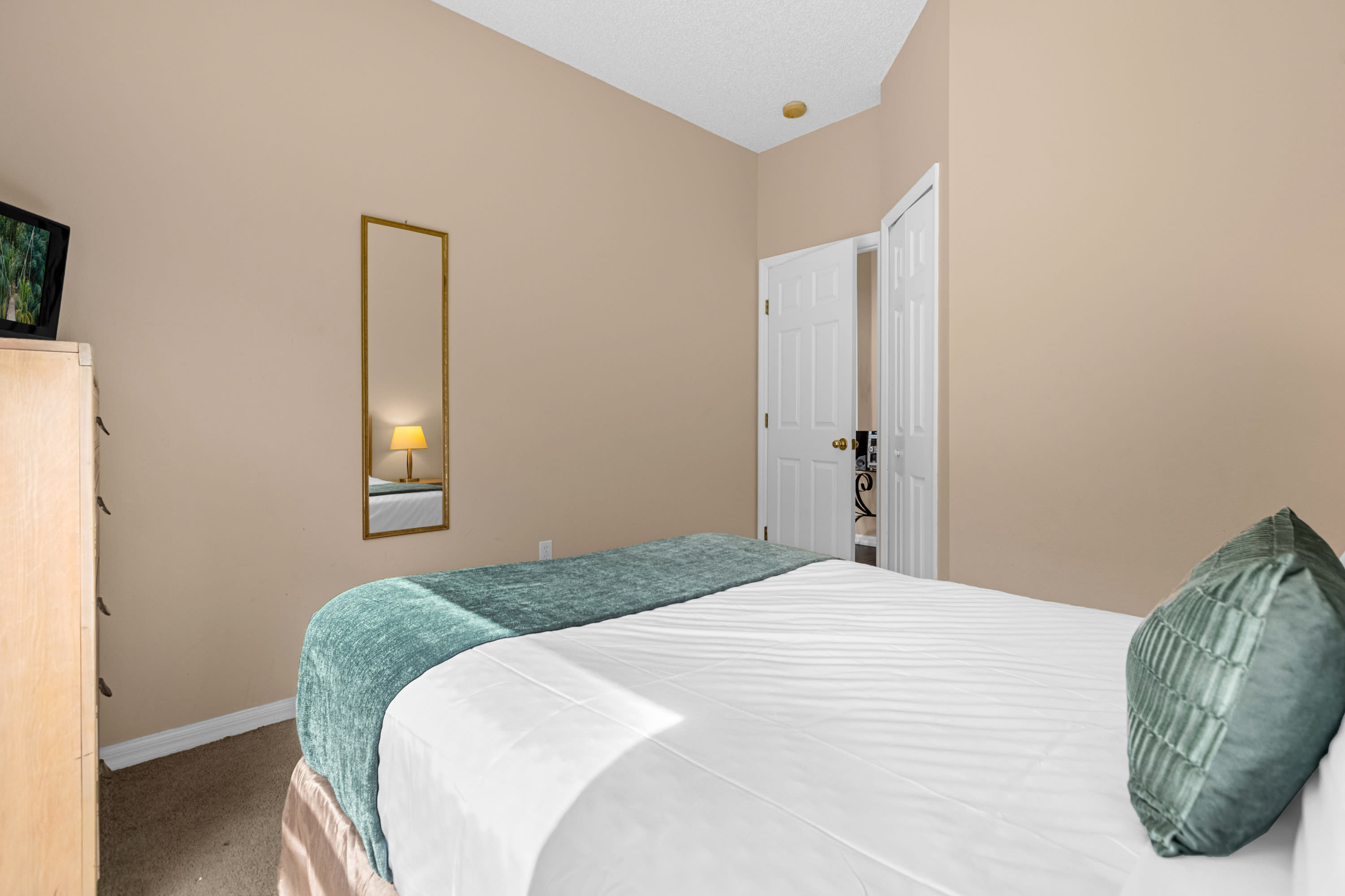 A different perspective of the bedroom, highlighting the queen bed with white bedding and a teal throw, and a full-length mirror on the wall. The room has light beige walls and a carpeted floor.