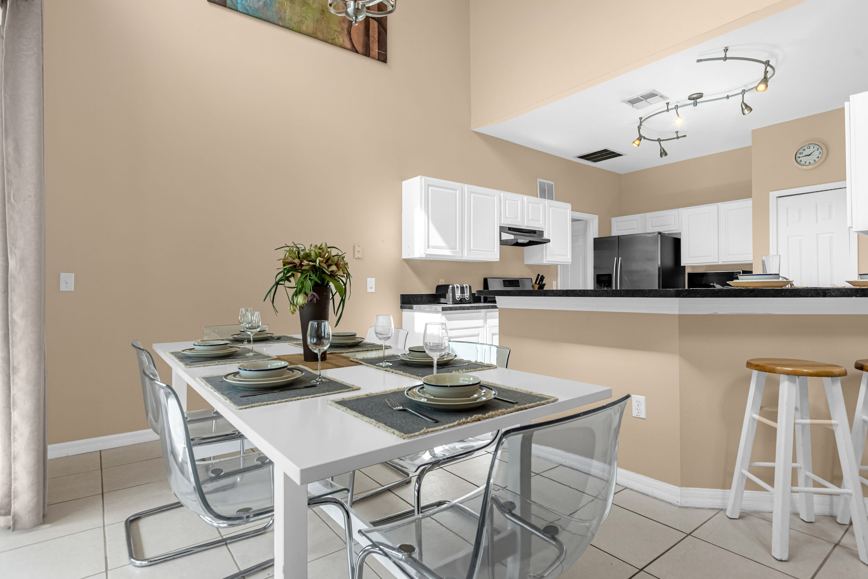 Modern dining area with a white rectangular table set for six, featuring clear acrylic chairs and place settings. It is situated next to a kitchen counter with a breakfast bar, under a chandelier.