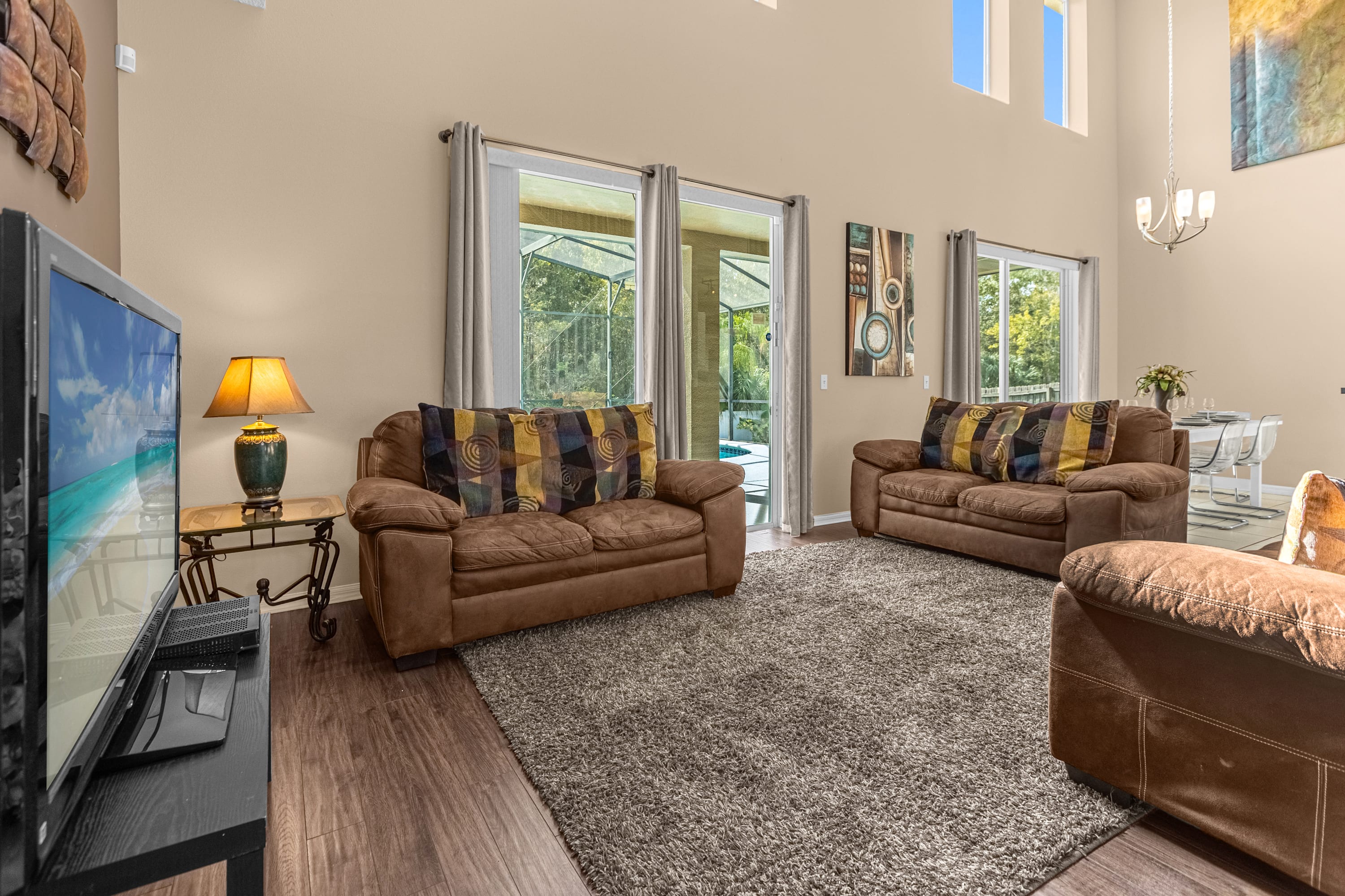 Bright living room featuring two brown sofas with patterned pillows, a flat-screen TV, and large sliding glass doors opening to a screened patio with a pool view. The room has high ceilings and light-colored walls.