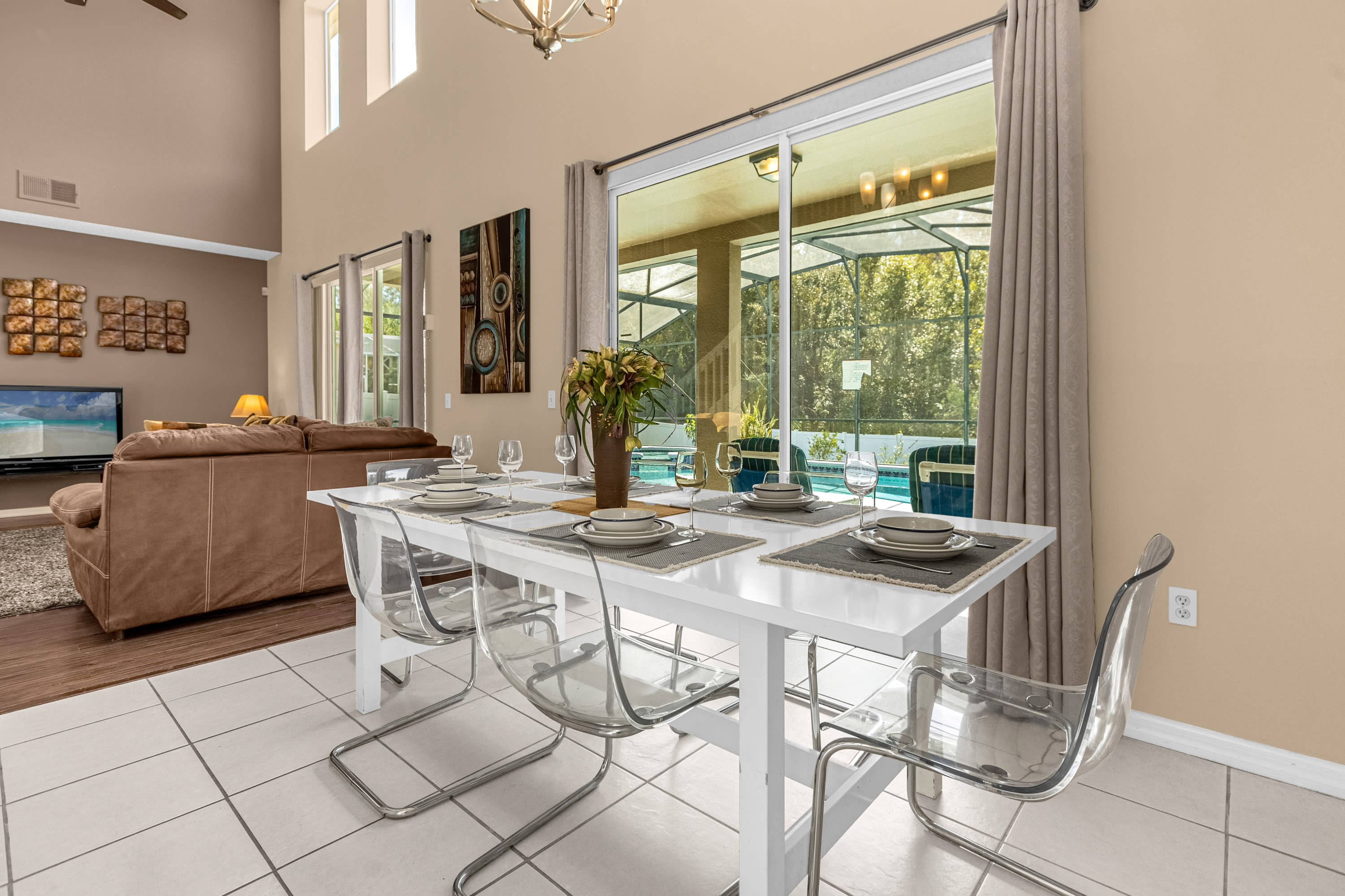 Modern dining area with a white rectangular table and clear acrylic chairs, set on white tiled flooring, with a view of the screened pool area through large sliding glass doors. The living room is visible in the background.