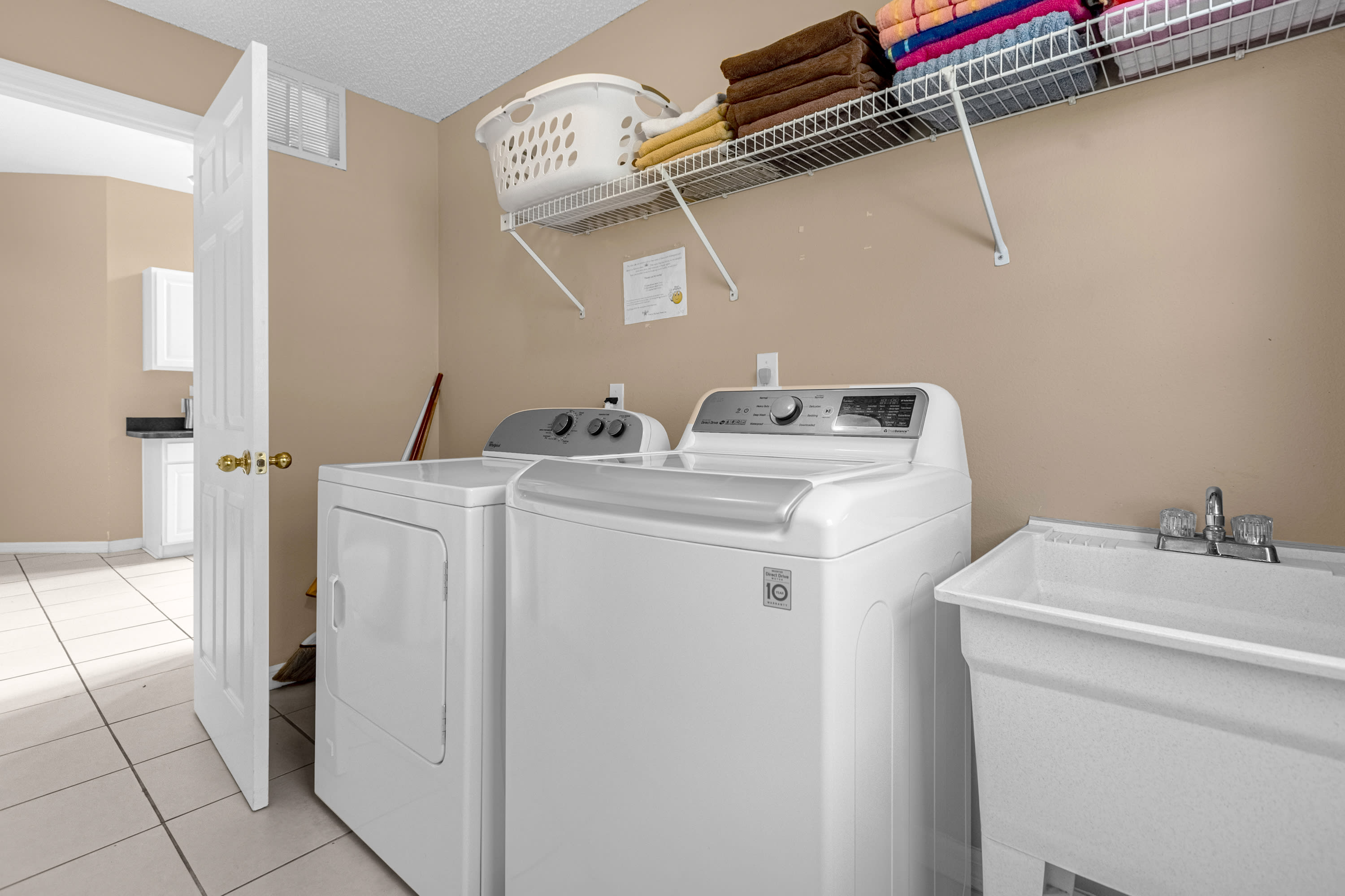 A dedicated laundry room equipped with a white top-loading washing machine and a front-loading dryer, a utility sink, and a wire shelf with towels and a laundry basket. The walls are light beige, and the floor is tiled.