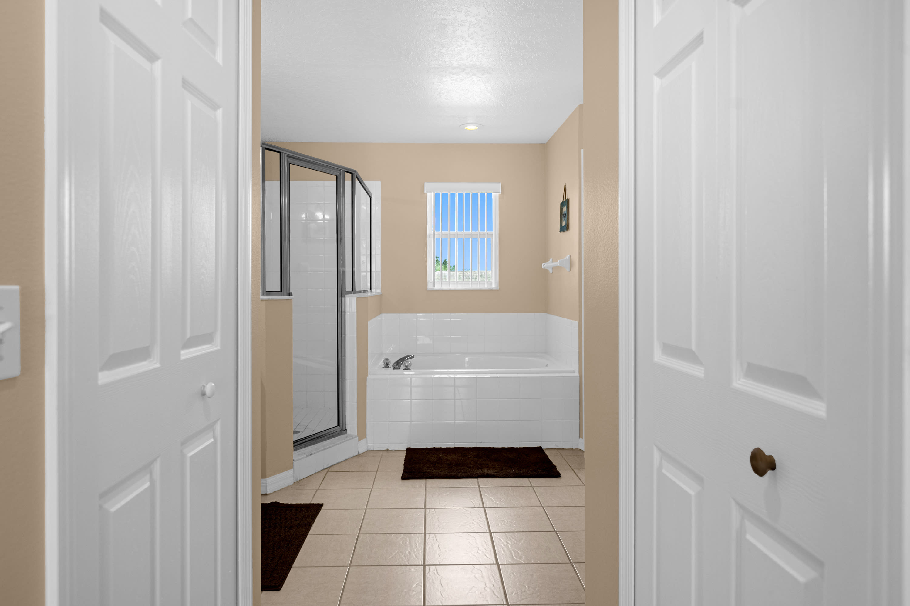 A view into a bathroom with a glass-enclosed walk-in shower and a white tiled bathtub under a window. The floor is tiled, and white doors are visible on either side of the entrance.