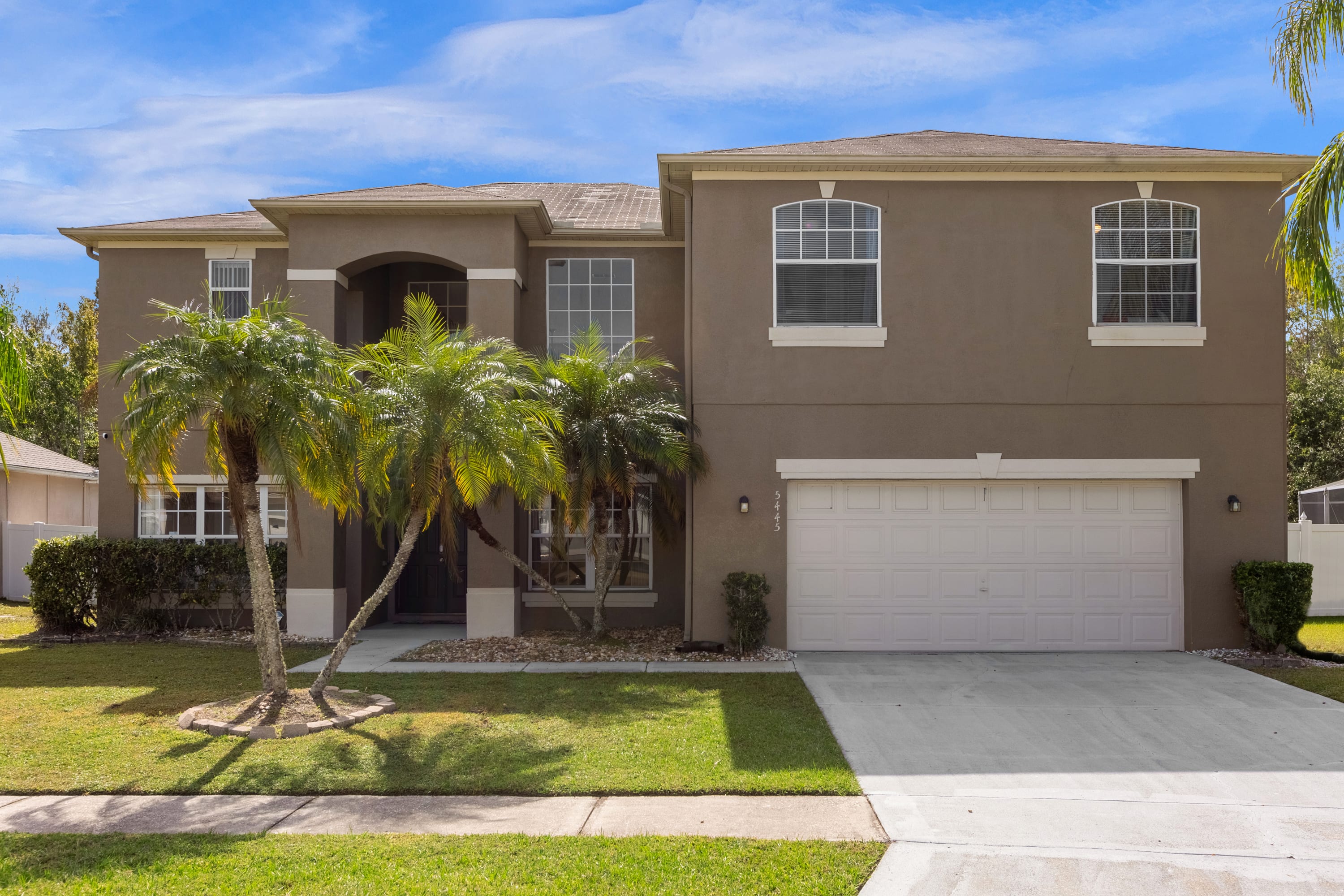 Two-story stucco house with a warm brown exterior, featuring a two-car garage, arched entryway, and palm trees in the front yard, under a clear blue sky.