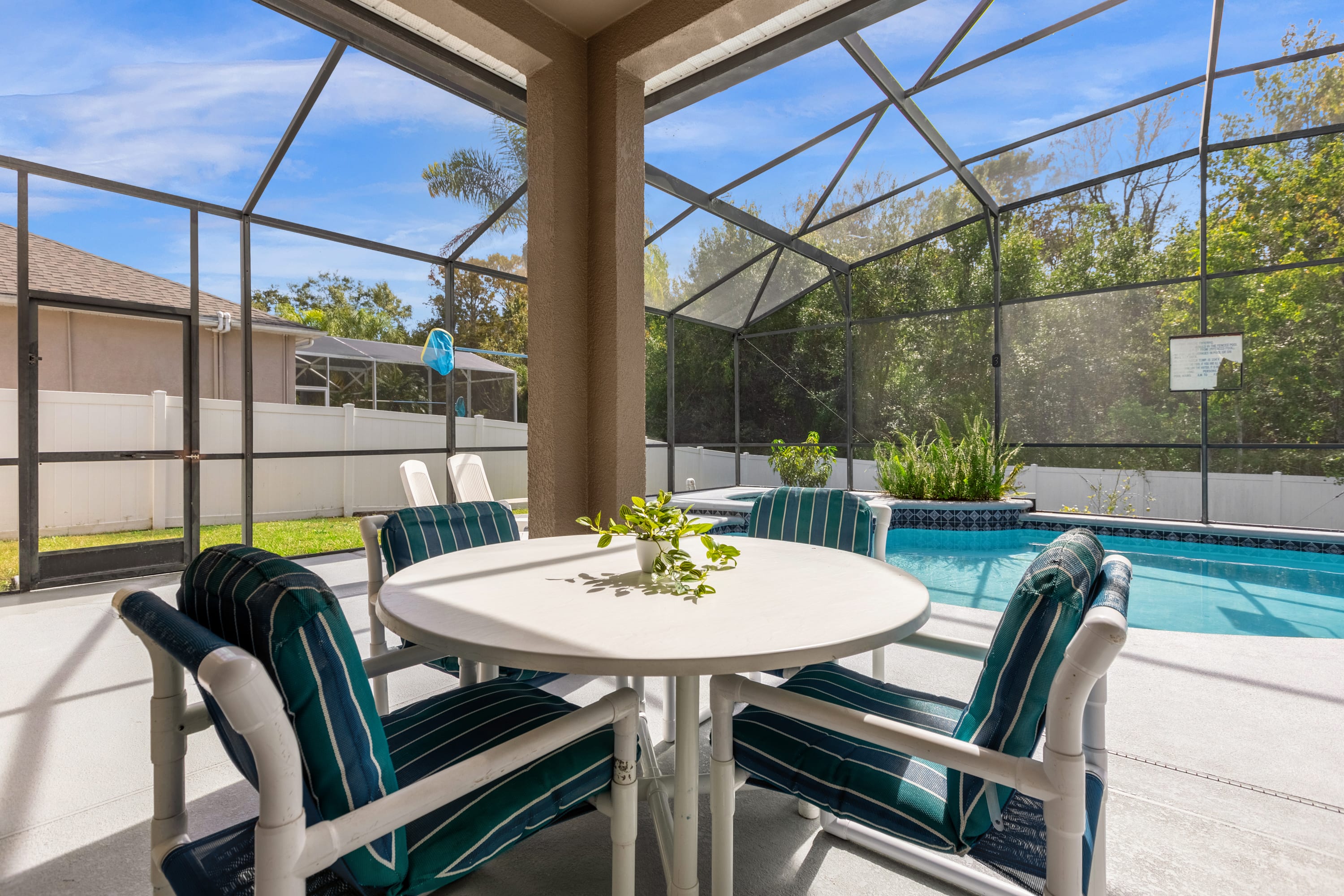 Screened-in patio area featuring a round white outdoor dining table with four chairs, offering a view of the swimming pool and lush greenery beyond the enclosure.