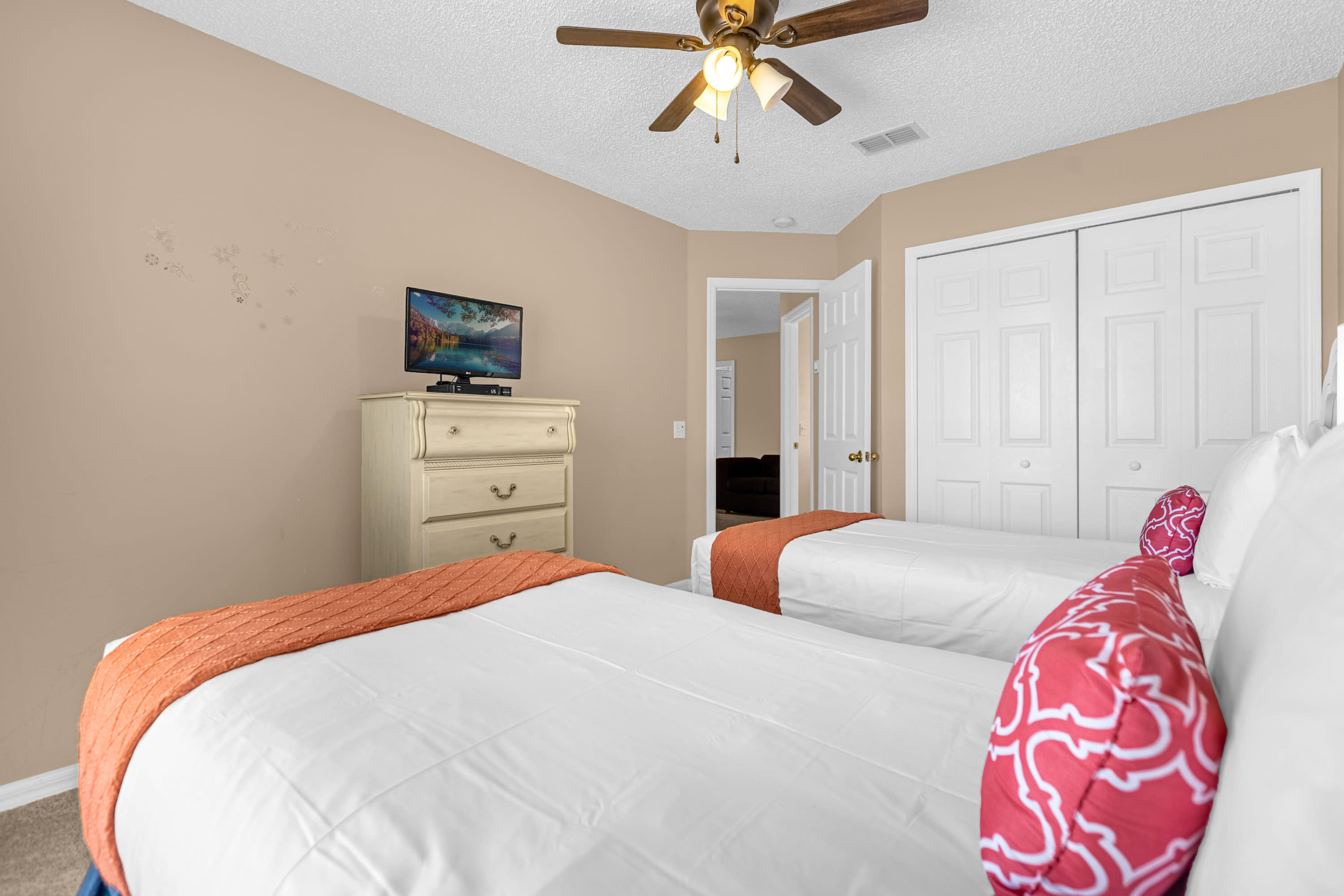 View of a bedroom with two twin beds, white bedding, and orange accents, showing a cream-colored dresser with a TV. A white bi-fold closet door is visible, and the room has light beige walls and carpet.