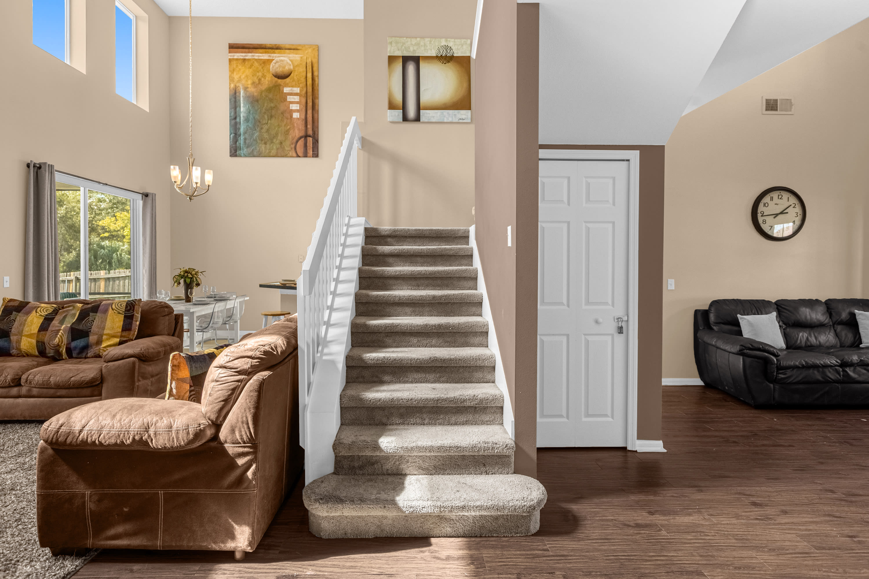 View of the carpeted staircase leading to the upper level, situated in an open-concept living space. The living room with brown sofas and the dining area are visible in the background, under high ceilings.