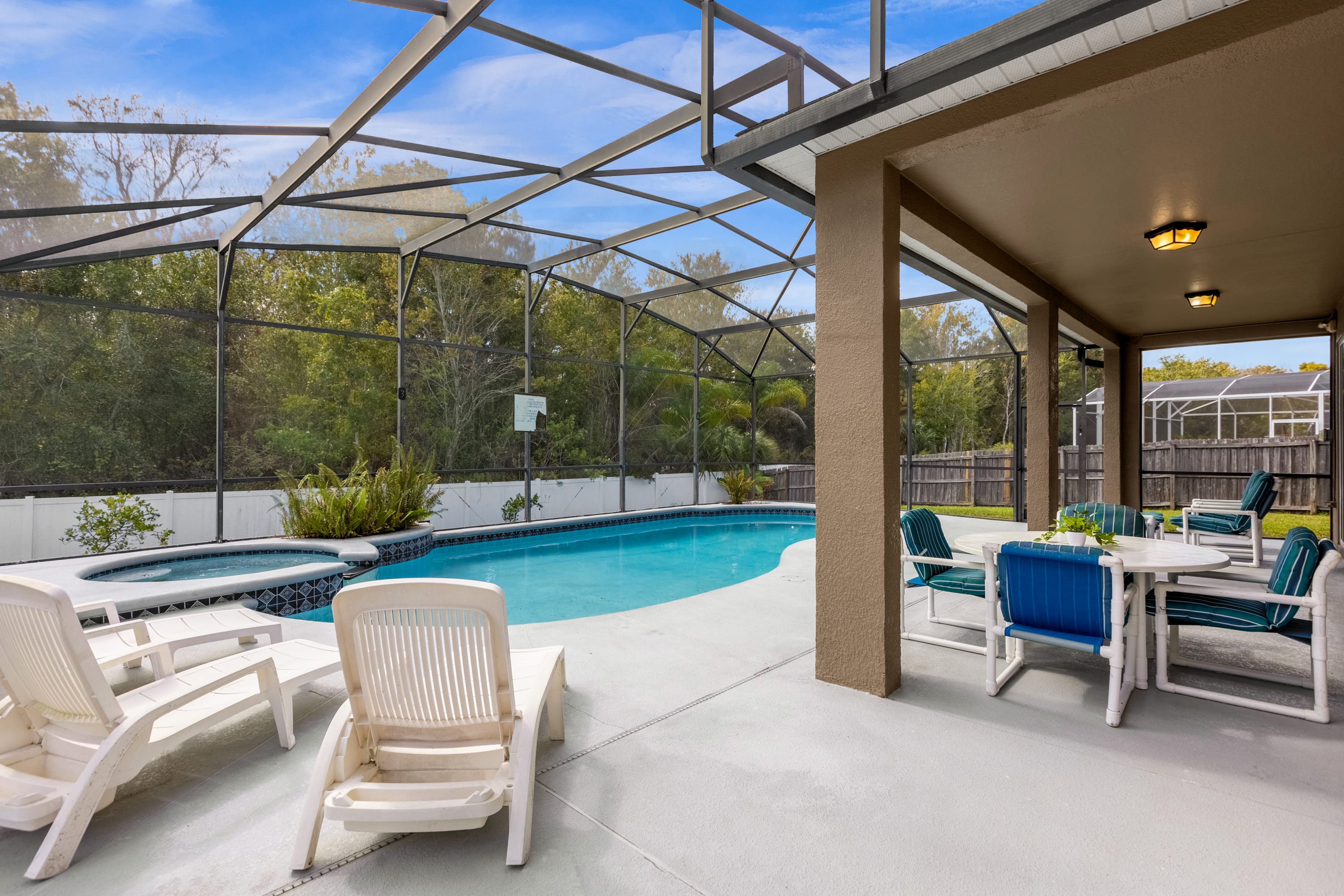 Covered patio area adjacent to the screened pool, furnished with white lounge chairs and an outdoor dining set, offering a shaded space overlooking the swimming pool and hot tub.