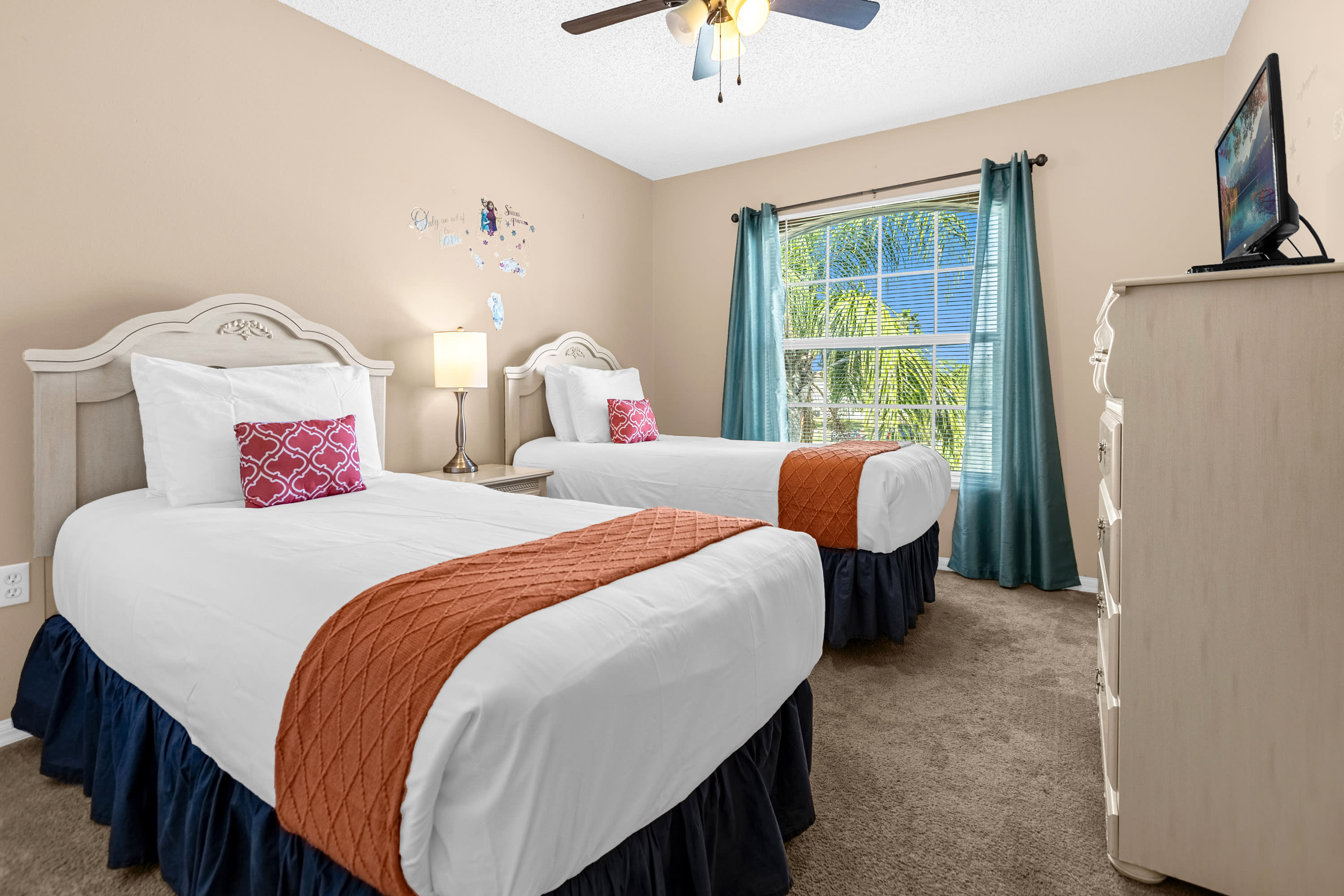 A bright bedroom with two twin beds featuring white headboards, white bedding, and orange throws, positioned under a large window with teal curtains. The room has light beige walls with wall decals and a ceiling fan.