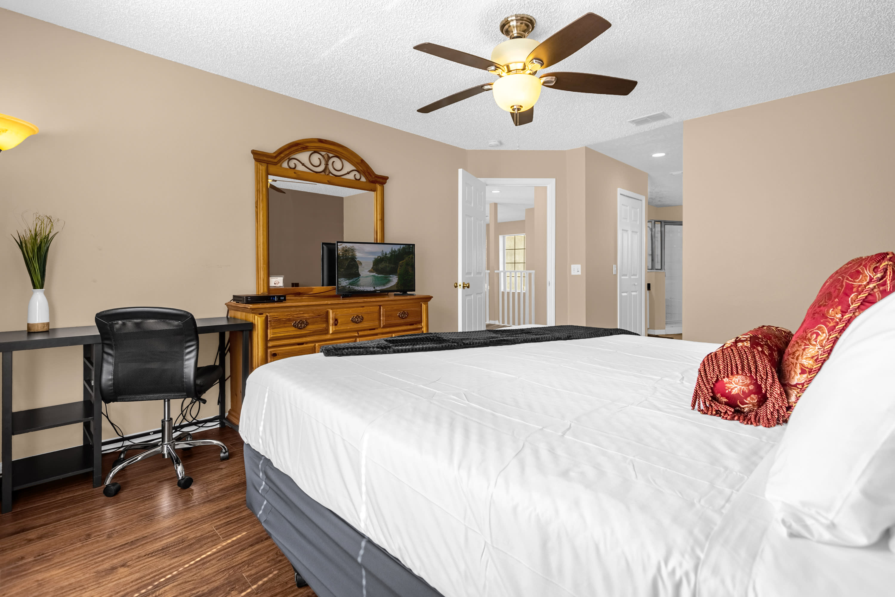 A spacious bedroom featuring a large bed with white bedding and red decorative pillows, a wooden dresser with a TV, and a black desk with an office chair. A ceiling fan with lights is visible overhead.
