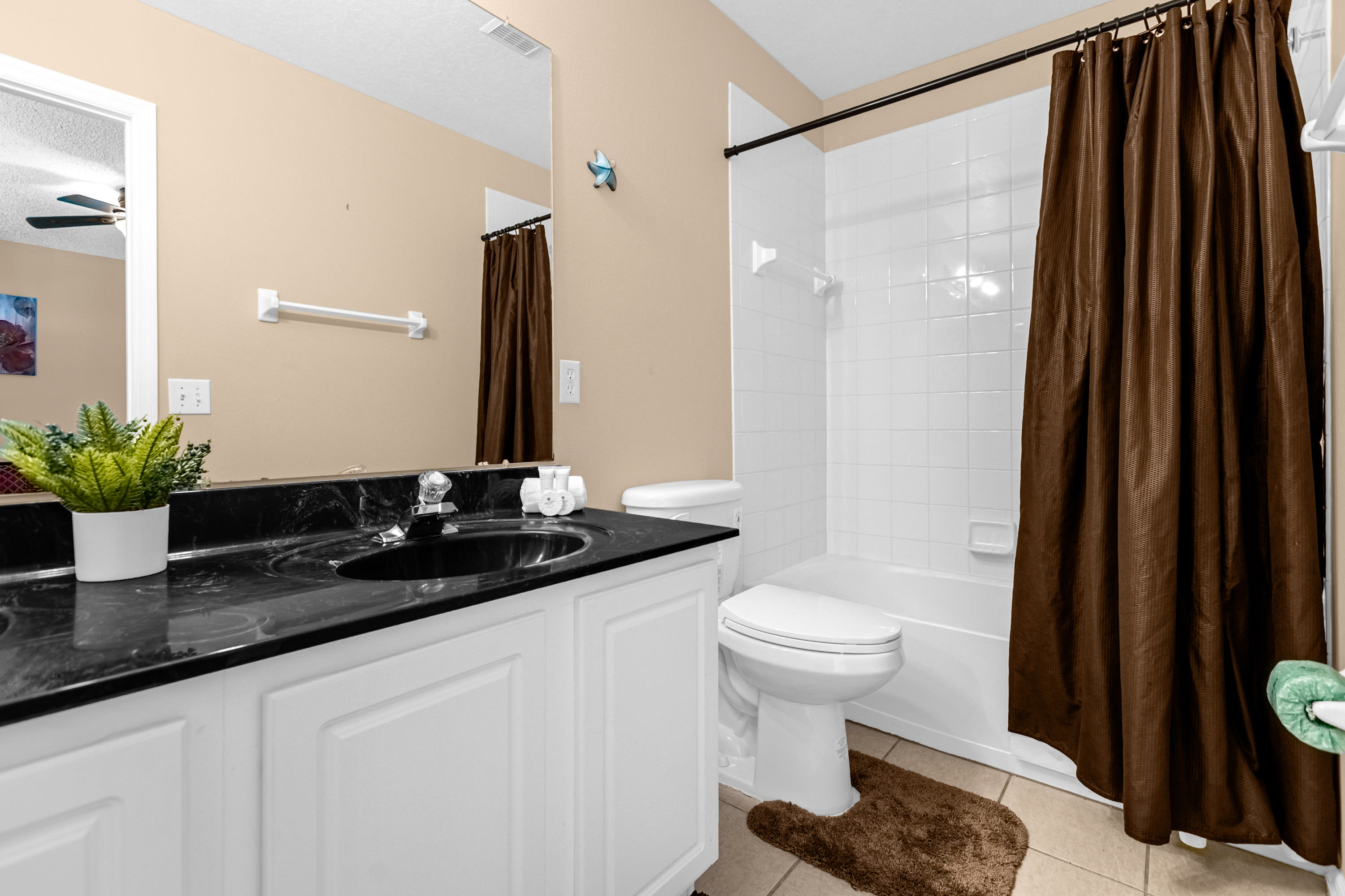 A functional bathroom with a white vanity, black countertop, and a single sink, featuring a toilet and a shower/tub combo with a dark brown shower curtain. The walls are light beige, and the floor is tiled.
