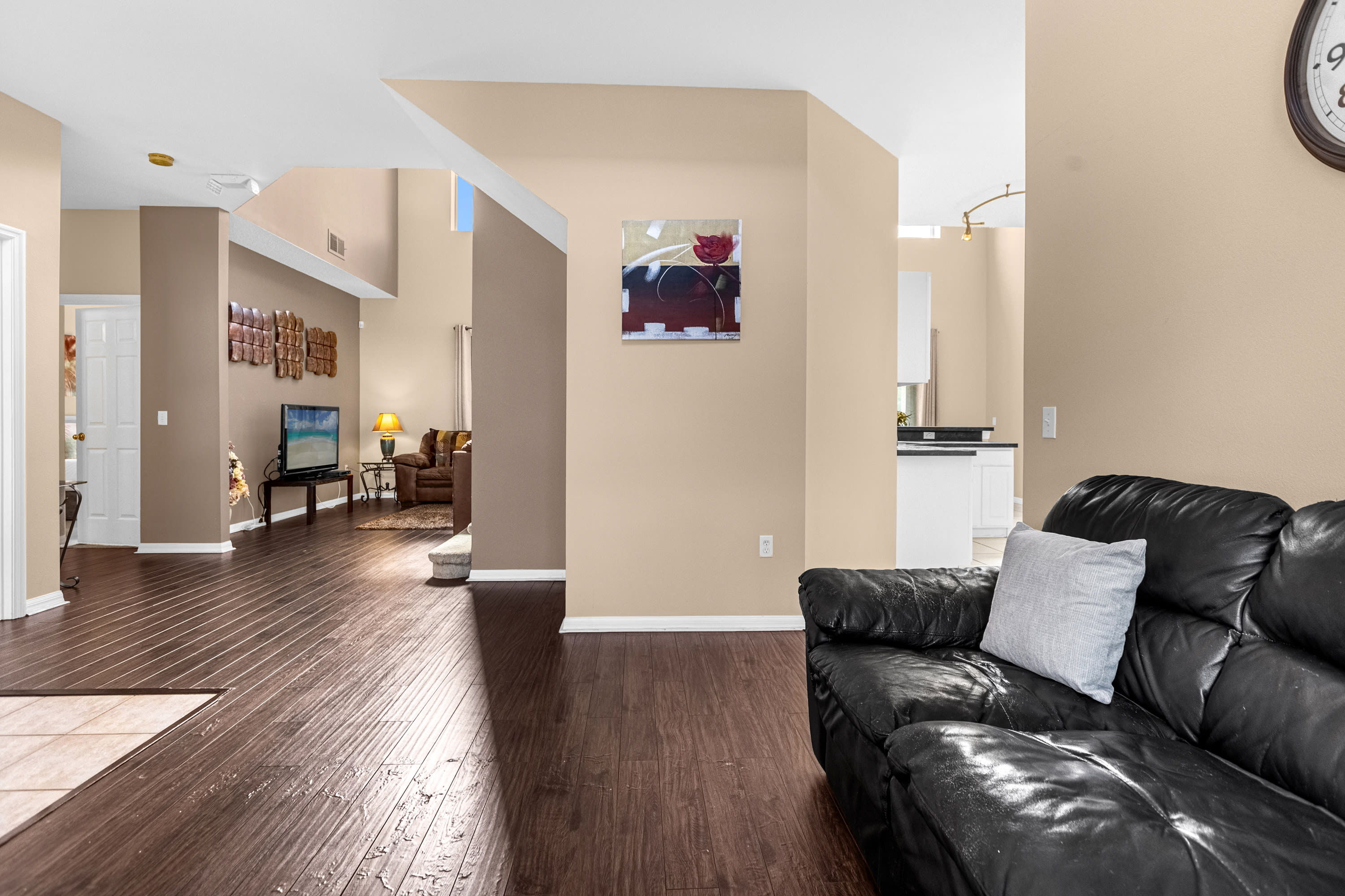 Transitional area with dark wood laminate flooring, connecting the living room and kitchen in an open-concept layout. A black leather sofa with a light pillow is visible, along with a wall adorned with artwork.
