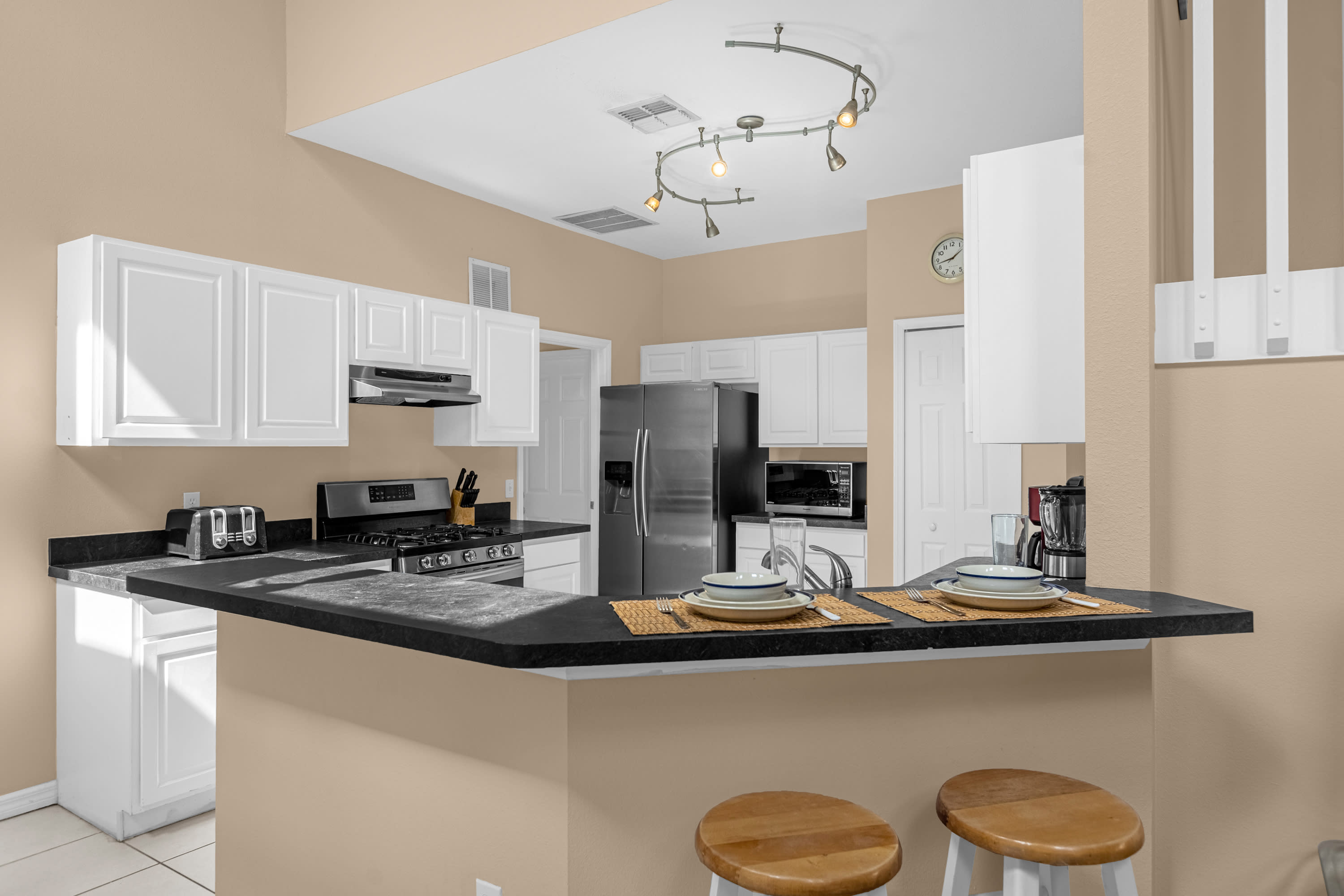 Kitchen view highlighting the breakfast bar with two wooden stools and the cooking area with a stainless steel gas range and microwave. White cabinets and dark countertops continue throughout, with a refrigerator visible in the background.