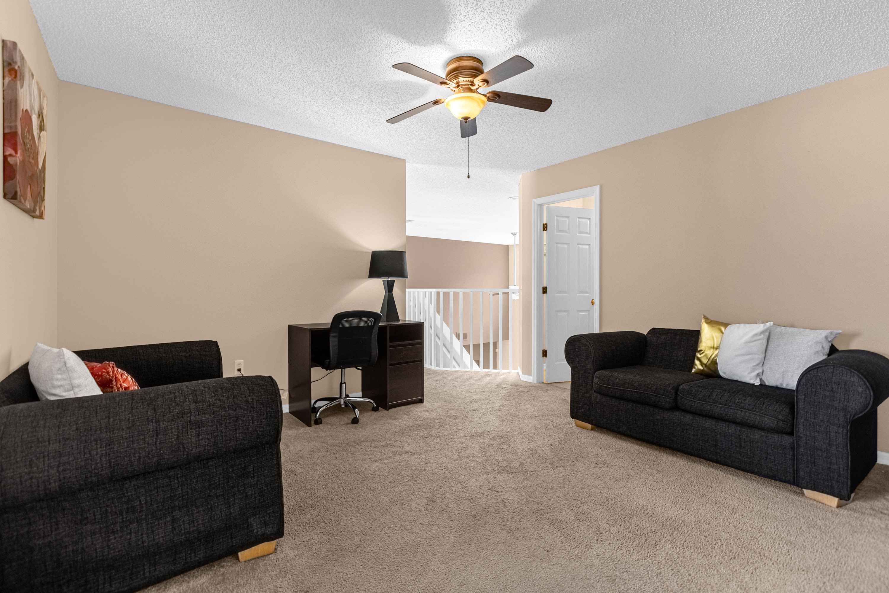 Another perspective of the upstairs loft, showcasing two dark sofas, a desk, and a railing overlooking the downstairs living area. The room is carpeted and features a ceiling fan and light-colored walls.