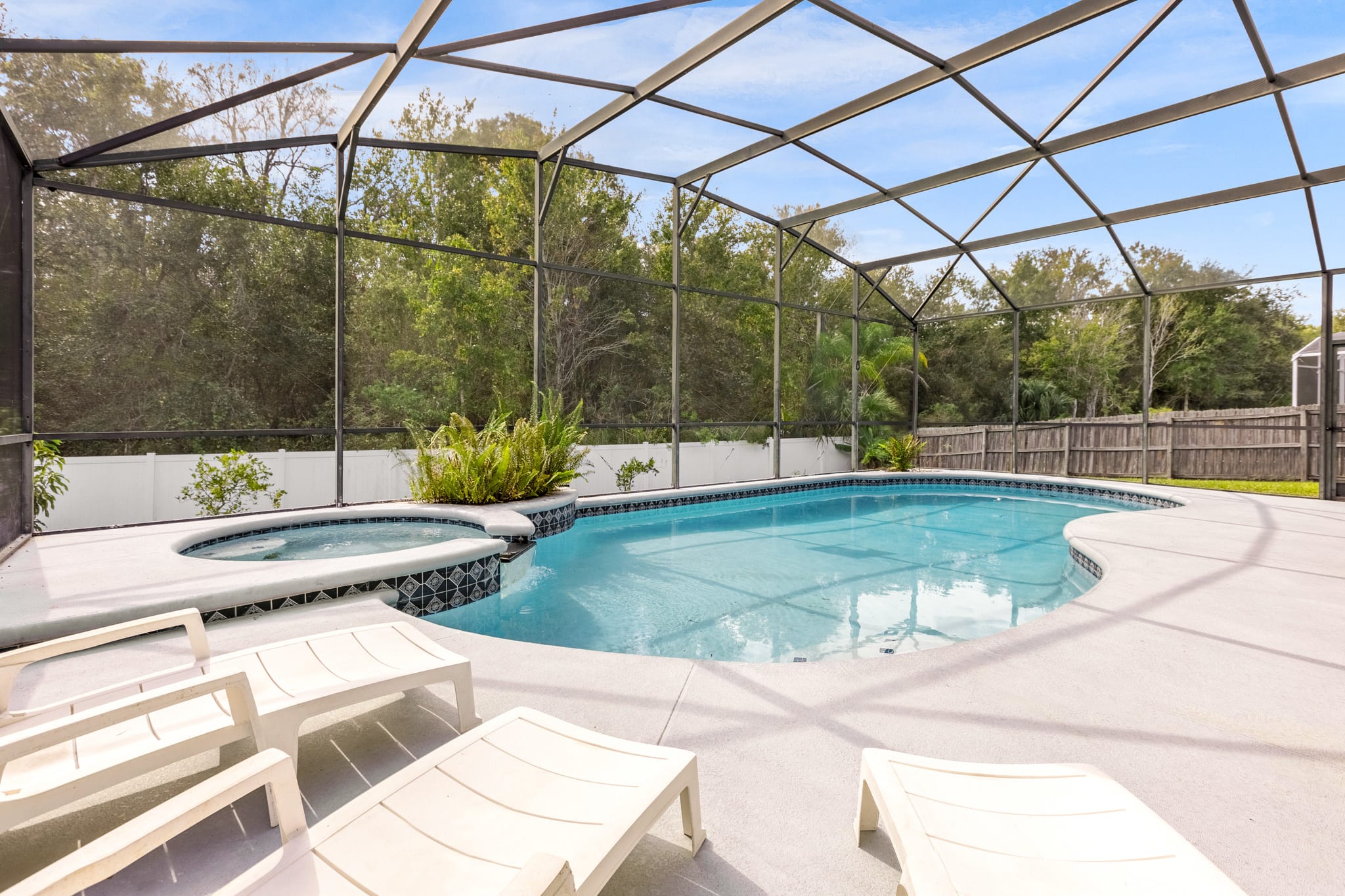 Expansive screened-in pool area with a freeform swimming pool and integrated hot tub, surrounded by a light concrete deck with several white lounge chairs, backed by a privacy fence and dense trees.