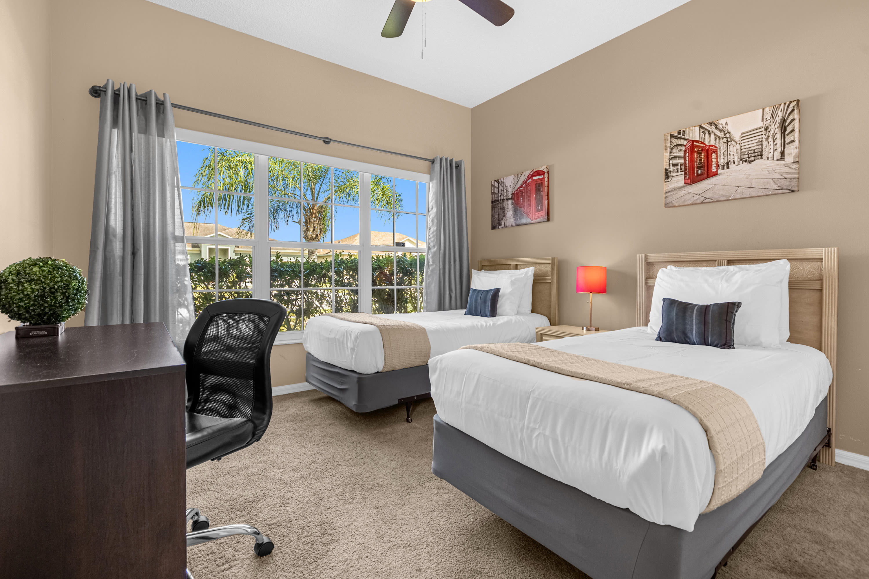 A twin bedroom with two beds featuring light wood headboards, white bedding, and beige throws, situated by a large window with gray curtains. A dark wood desk with an office chair and a red lamp on a nightstand are also visible.