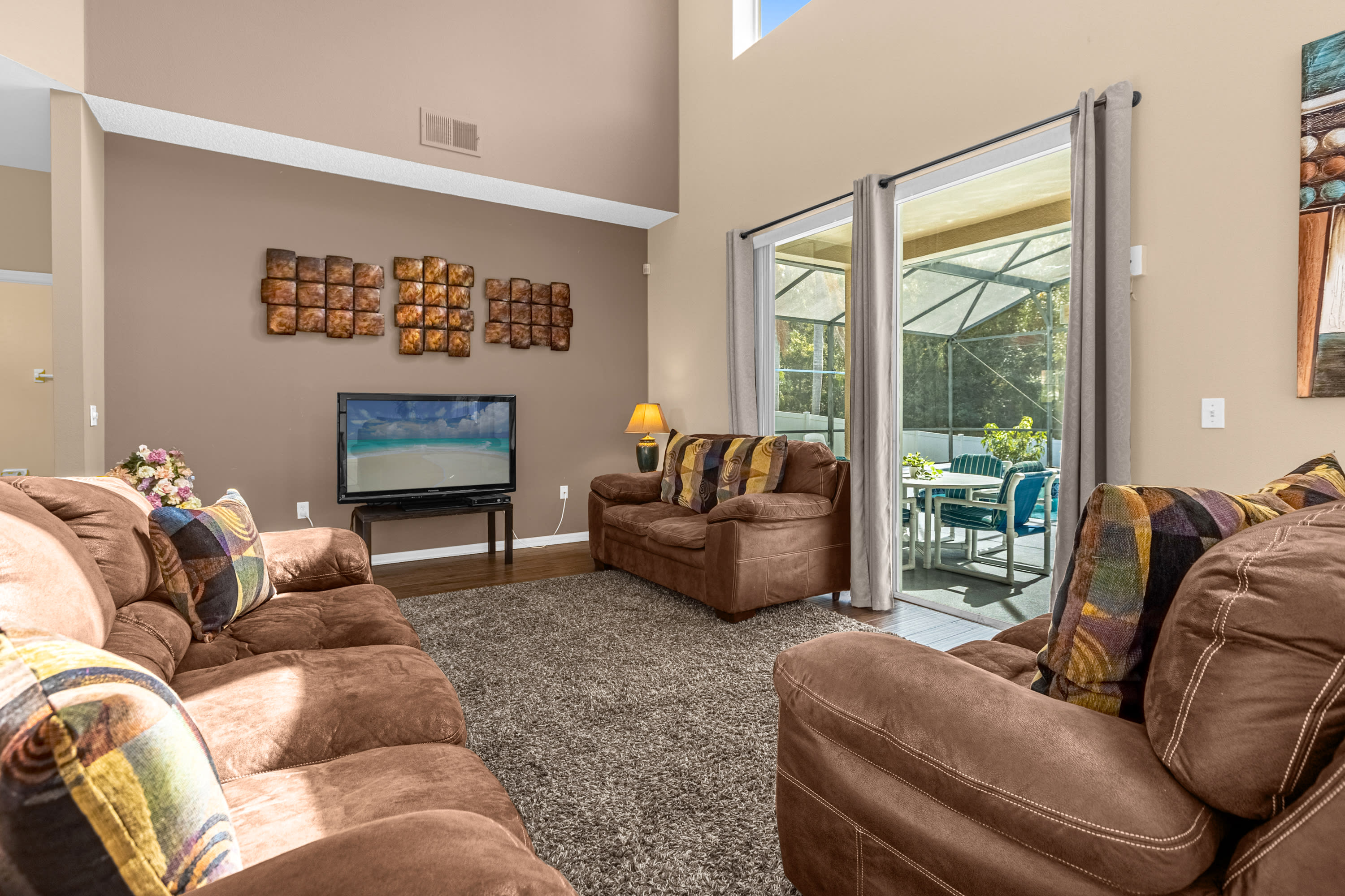 Spacious living room with a large brown sectional sofa, a flat-screen TV on a stand, and sliding glass doors leading to a screened outdoor area. The walls are a warm beige, and the floor is a mix of wood laminate and a large area rug.