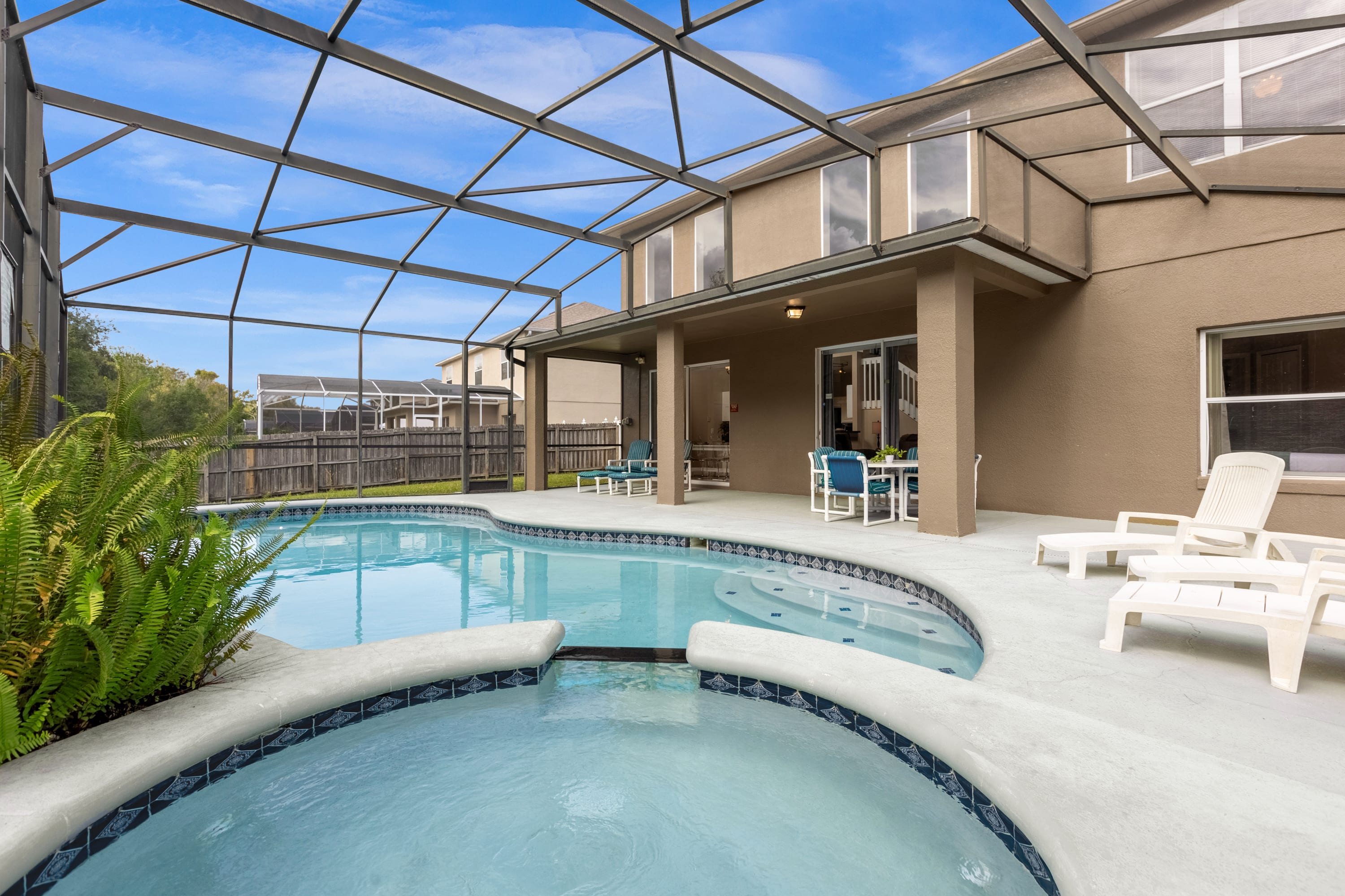 Rear view of the two-story house with a screened-in pool area, featuring a freeform pool and hot tub, a covered patio, and lounge chairs, all surrounded by a wooden fence and greenery.