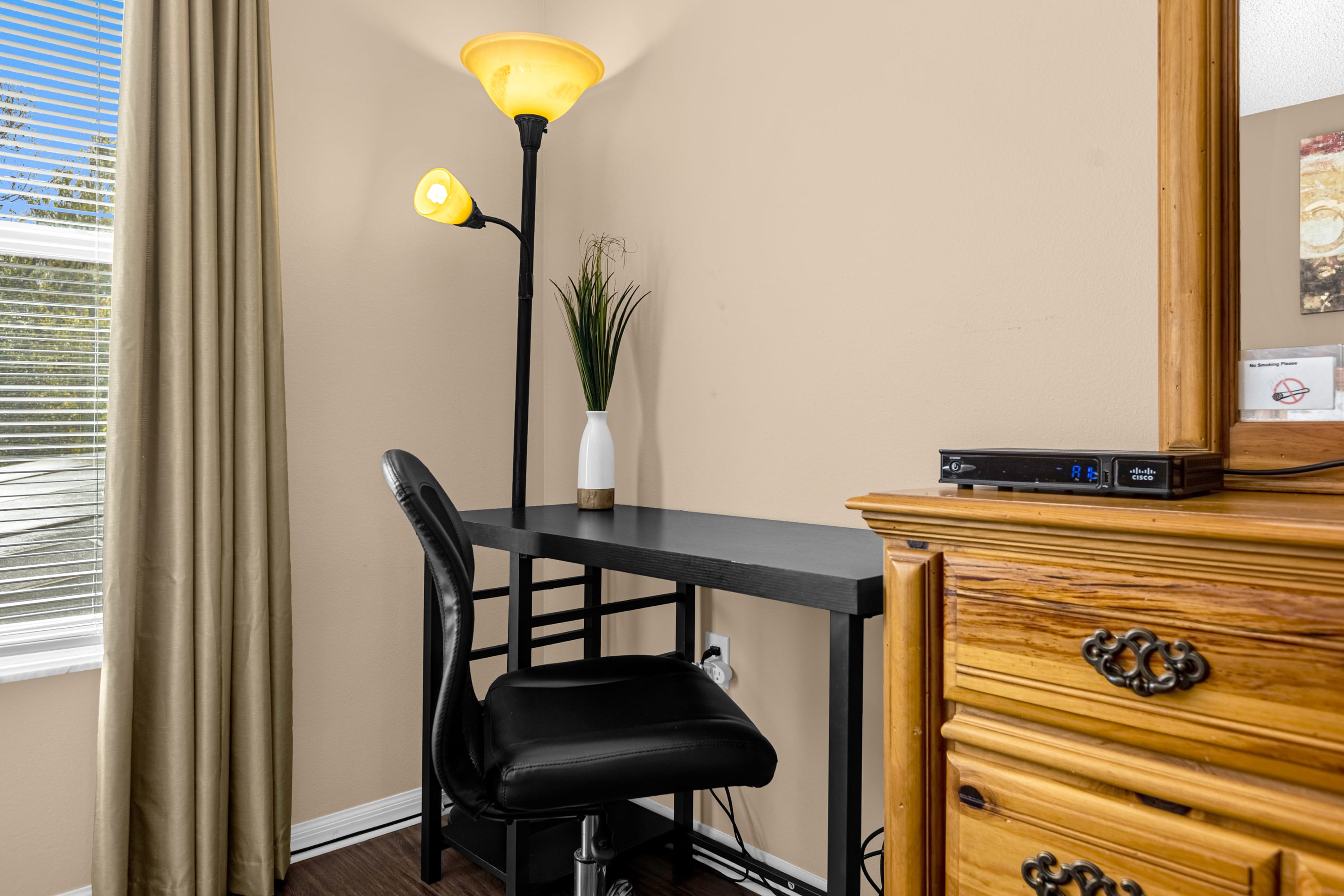 A corner of a bedroom with a black desk, a black office chair, and a floor lamp with two yellow shades. A white vase with greenery sits on the desk next to a window with blinds.