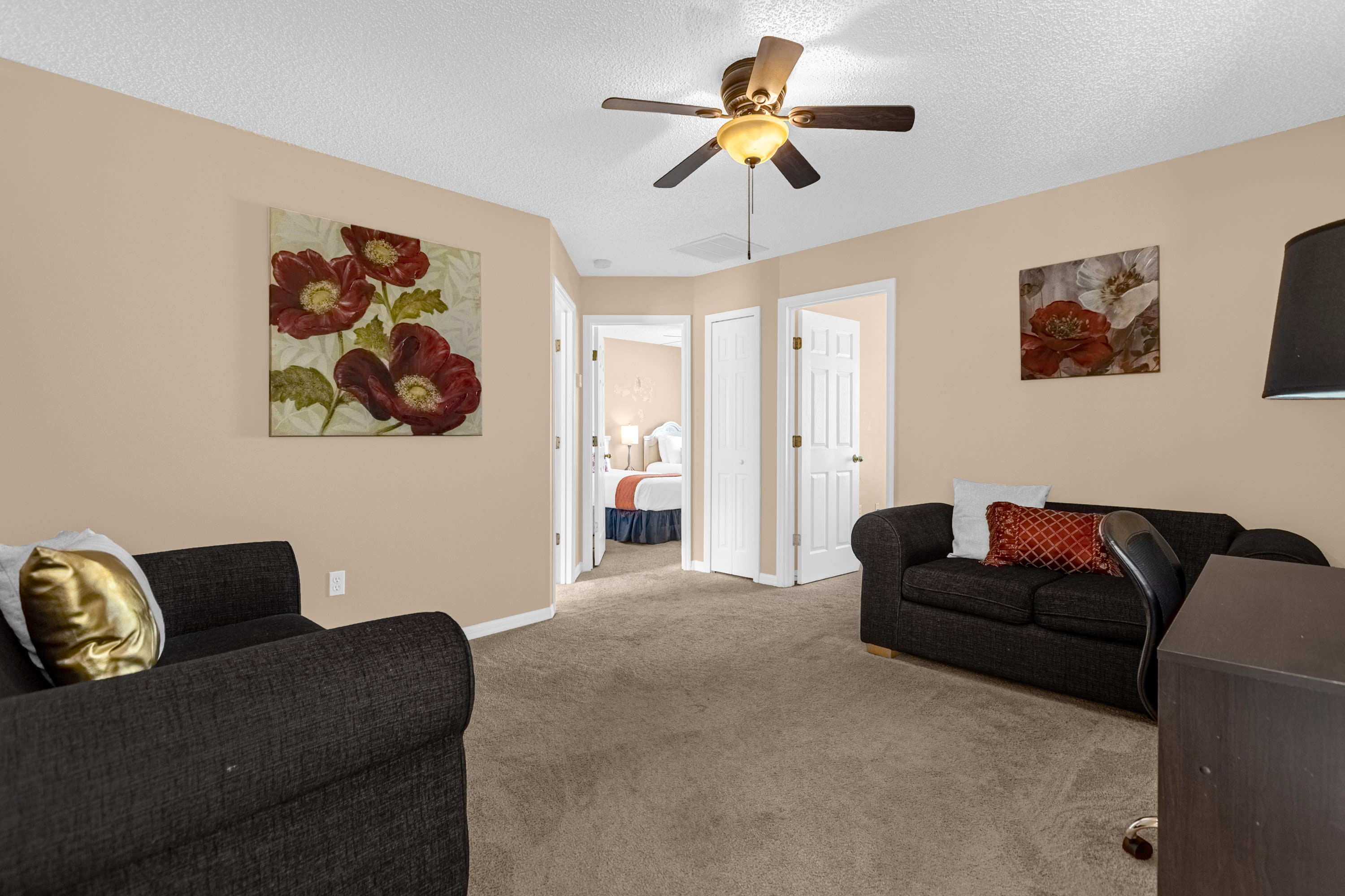 Upstairs loft area furnished with two dark sofas and a desk with a chair, featuring carpeted flooring and light beige walls. Artwork adorns the walls, and a ceiling fan provides air circulation.