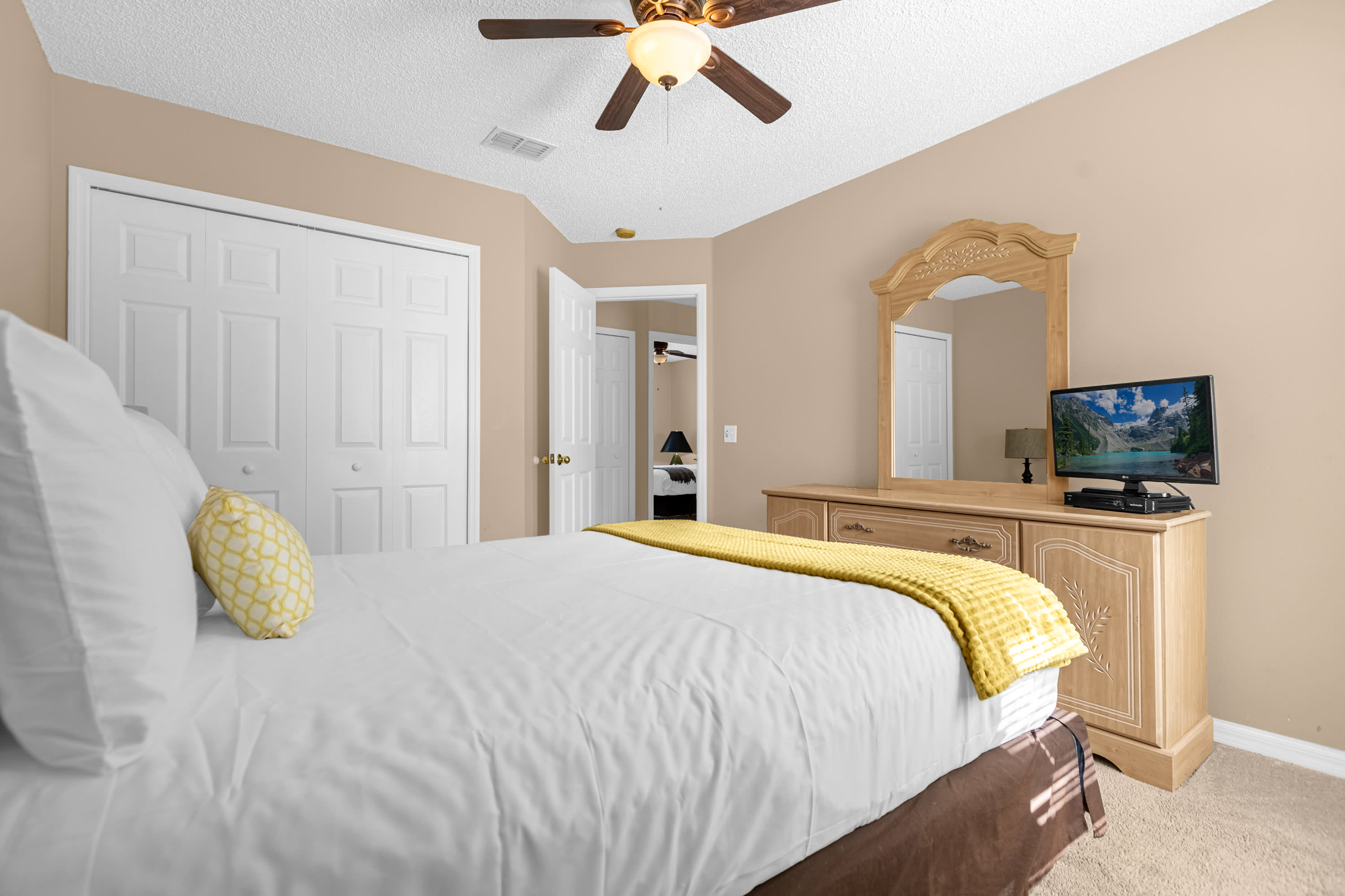 A different angle of the bedroom, showing the queen bed with white bedding and a yellow throw, a wooden dresser with a TV, and white bi-fold closet doors. A ceiling fan is visible overhead.