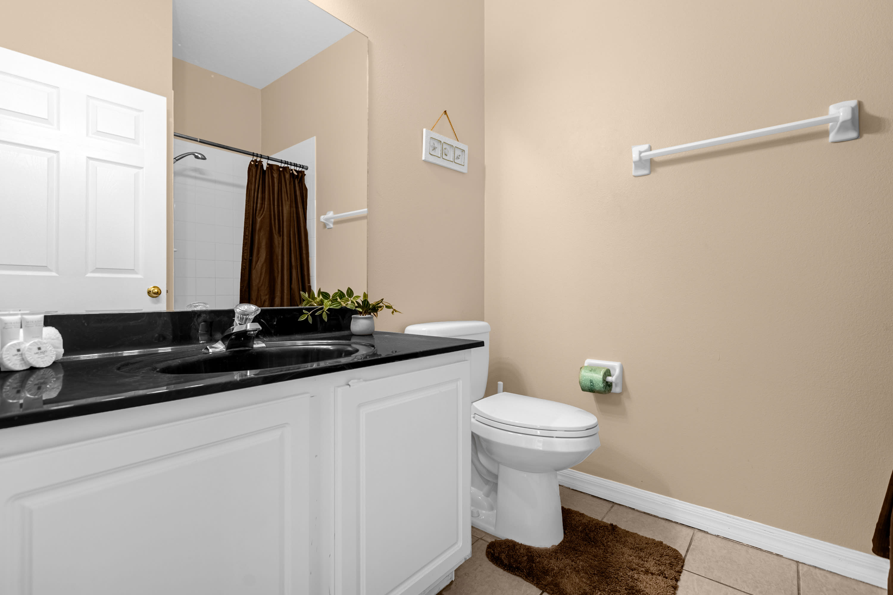 A compact bathroom featuring a white vanity with a black countertop and single sink, a toilet, and a mirror. A brown bath mat is on the tiled floor, and a shower/tub combo with a brown curtain is partially visible.