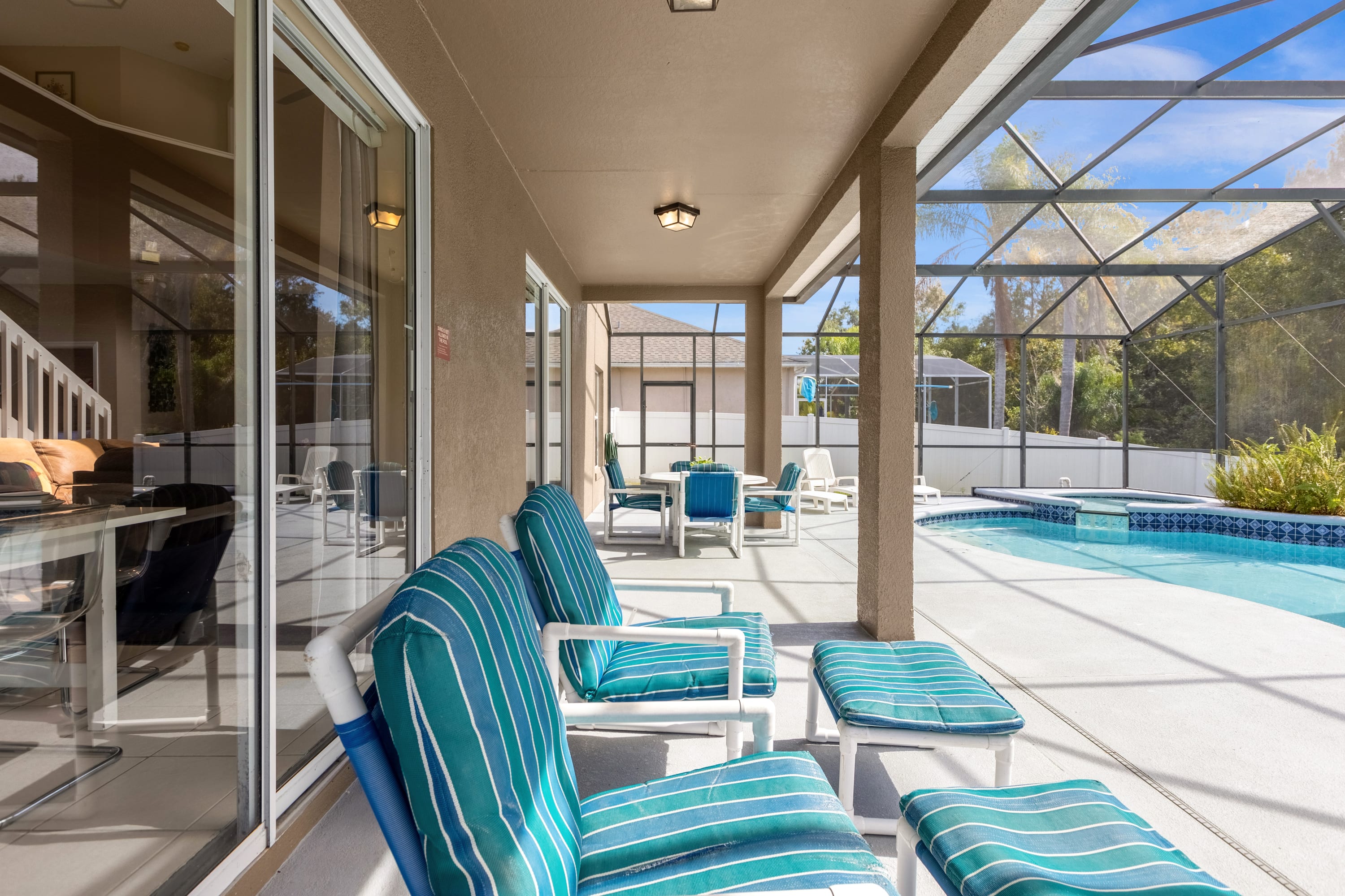 Covered patio area with striped lounge chairs and an outdoor dining set, positioned next to the screened pool, with large sliding glass doors providing access to the interior of the house.