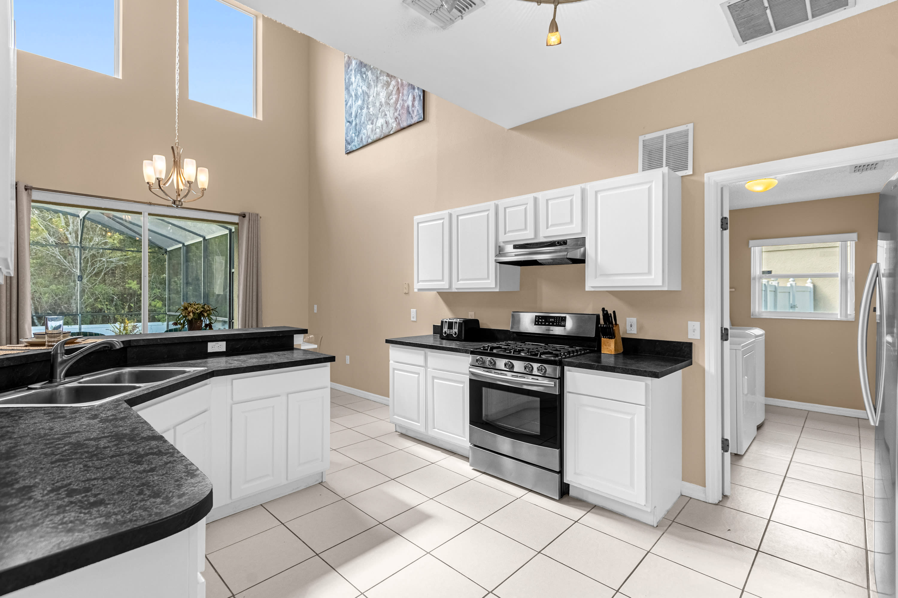 Expansive kitchen featuring white cabinetry, dark countertops, and stainless steel appliances, including a gas range and double sink. The room has white tile flooring and an open layout, with a doorway leading to a laundry area.