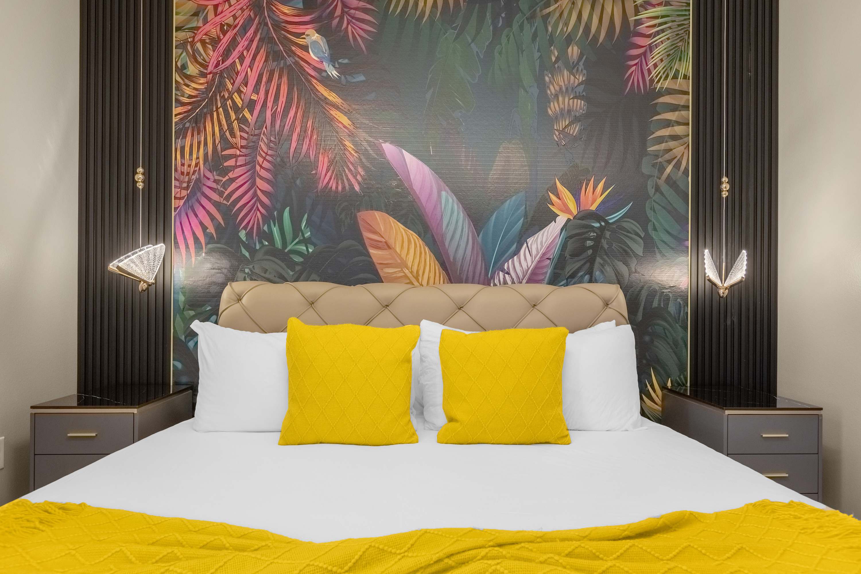 Close-up view of a king-sized bed with a beige upholstered headboard, adorned with white and bright yellow decorative pillows and a textured yellow throw blanket. The vibrant tropical mural and unique hanging lights on the accent wall are prominent.