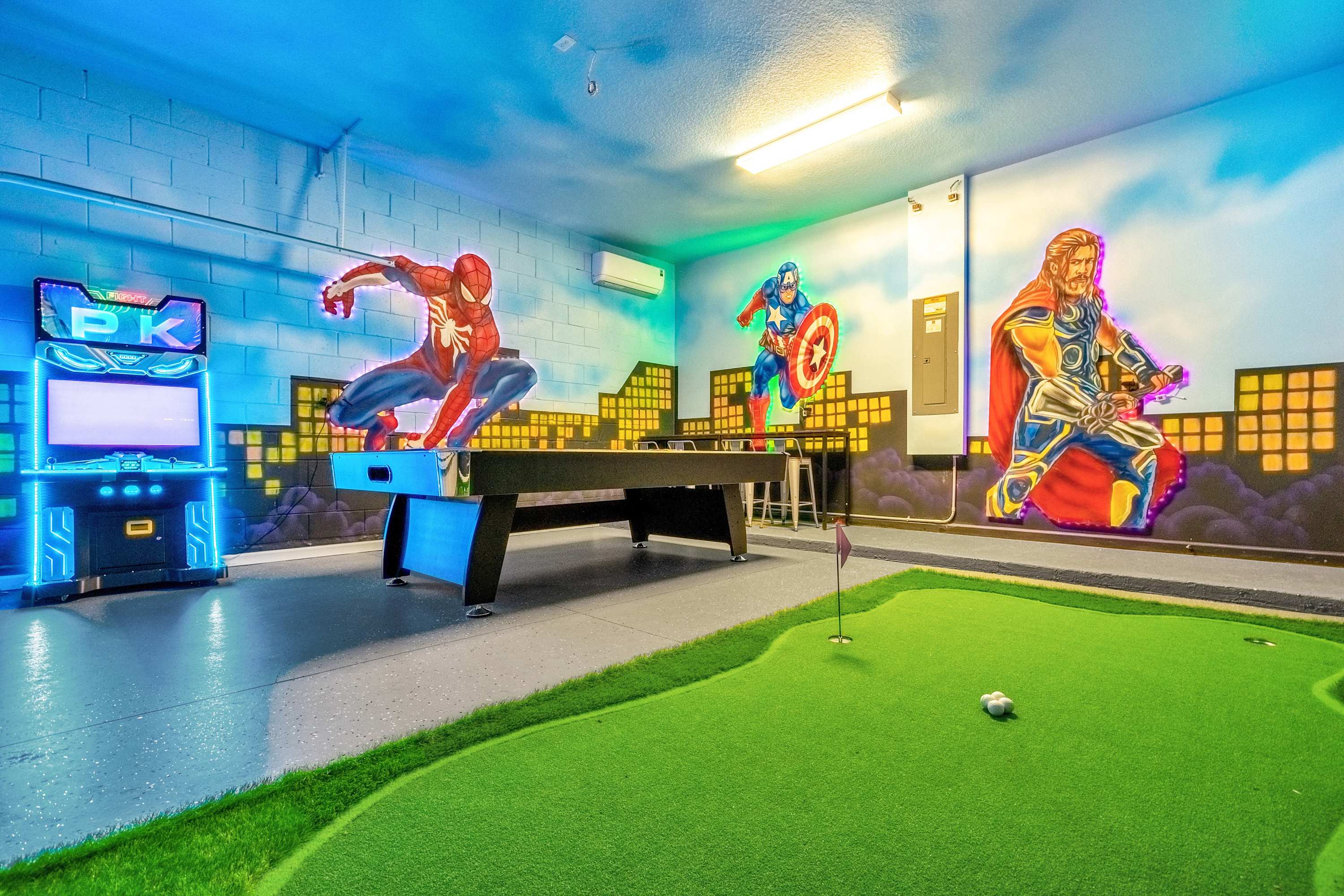 A vibrant themed game room features a pool table and a putting green, set against walls adorned with colorful superhero murals and neon lighting. An arcade game machine with glowing accents adds to the entertainment options.