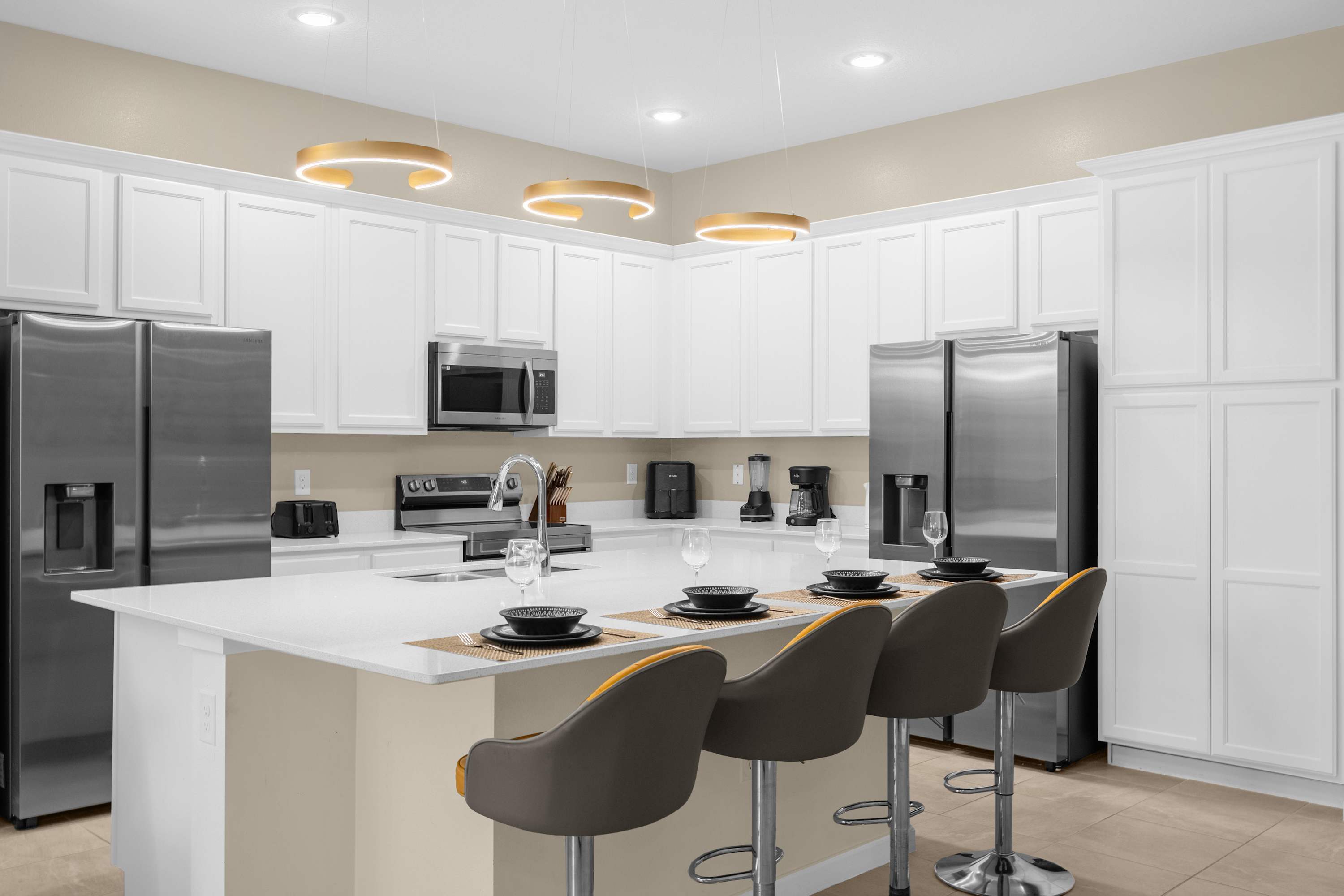 A bright, modern kitchen featuring white shaker cabinets, stainless steel appliances including two refrigerators, and a large white quartz island with a sink and bar seating. Contemporary gold-ringed pendant lights illuminate the island.