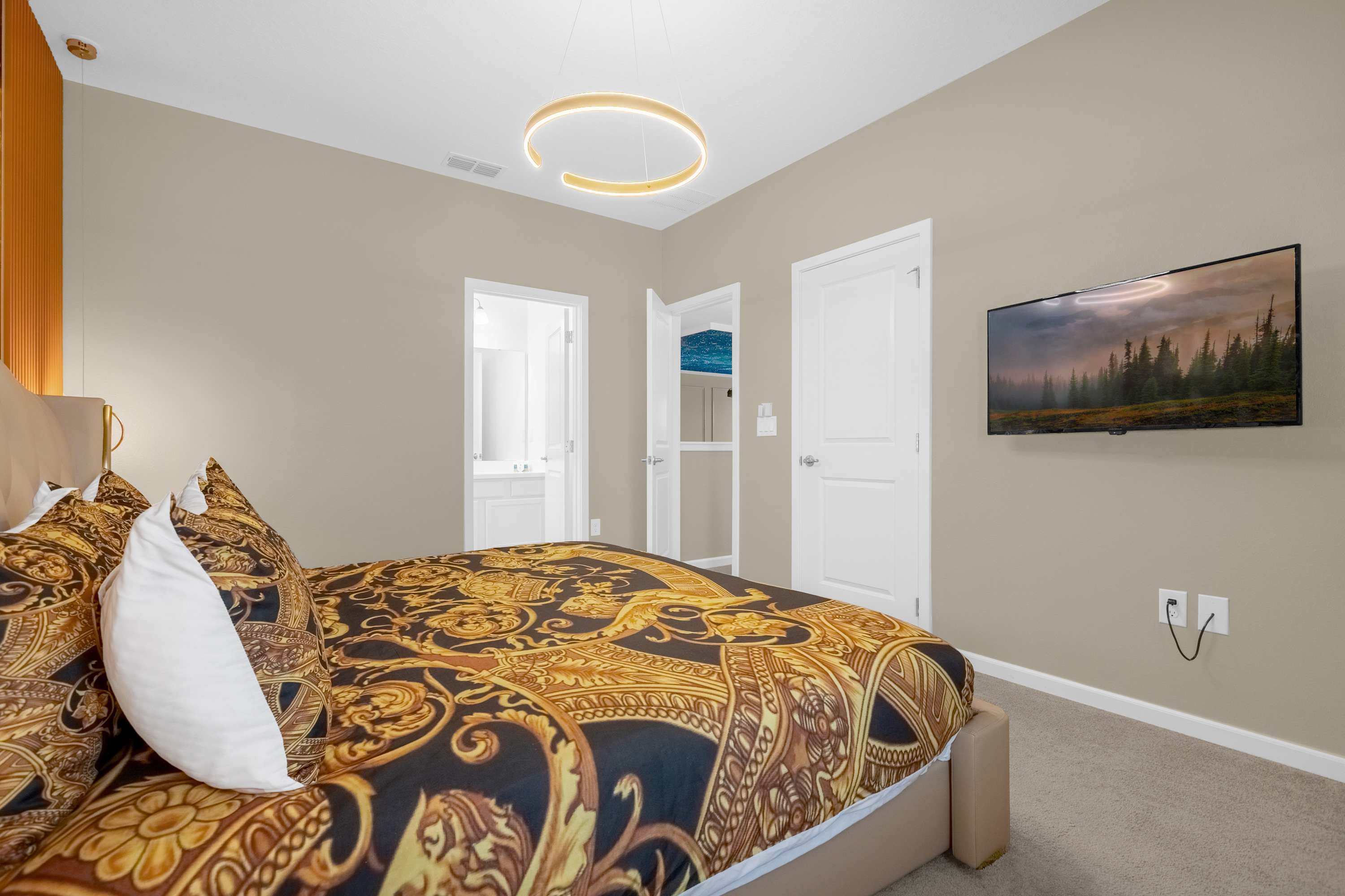 View of a lavish bedroom with a king-sized bed featuring black and gold patterned bedding, facing a wall-mounted flat-screen TV. A doorway leads to an en-suite bathroom, and white closet doors are visible.