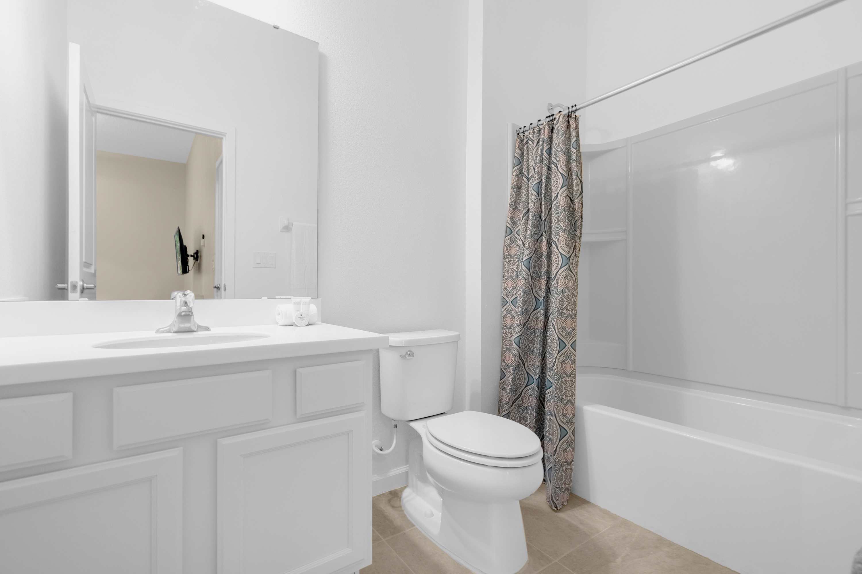Clean white bathroom featuring a single vanity with a mirror, a toilet, and a shower-tub combo with a patterned shower curtain, all set on light-colored tiled flooring.