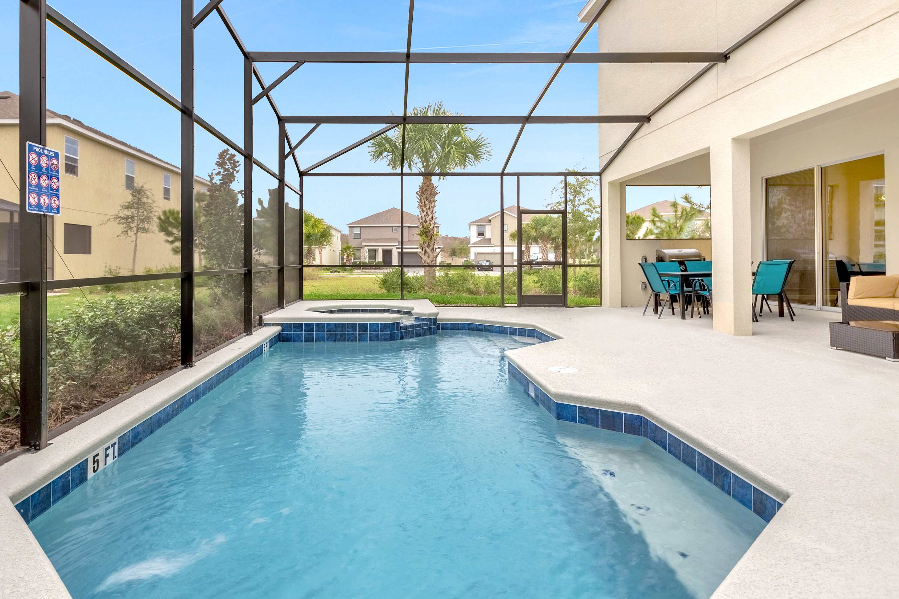 A spacious screened-in outdoor pool area features a large swimming pool with a connected hot tub, surrounded by a light-colored concrete deck. Beyond the screen, other houses and palm trees are visible under a clear blue sky.
