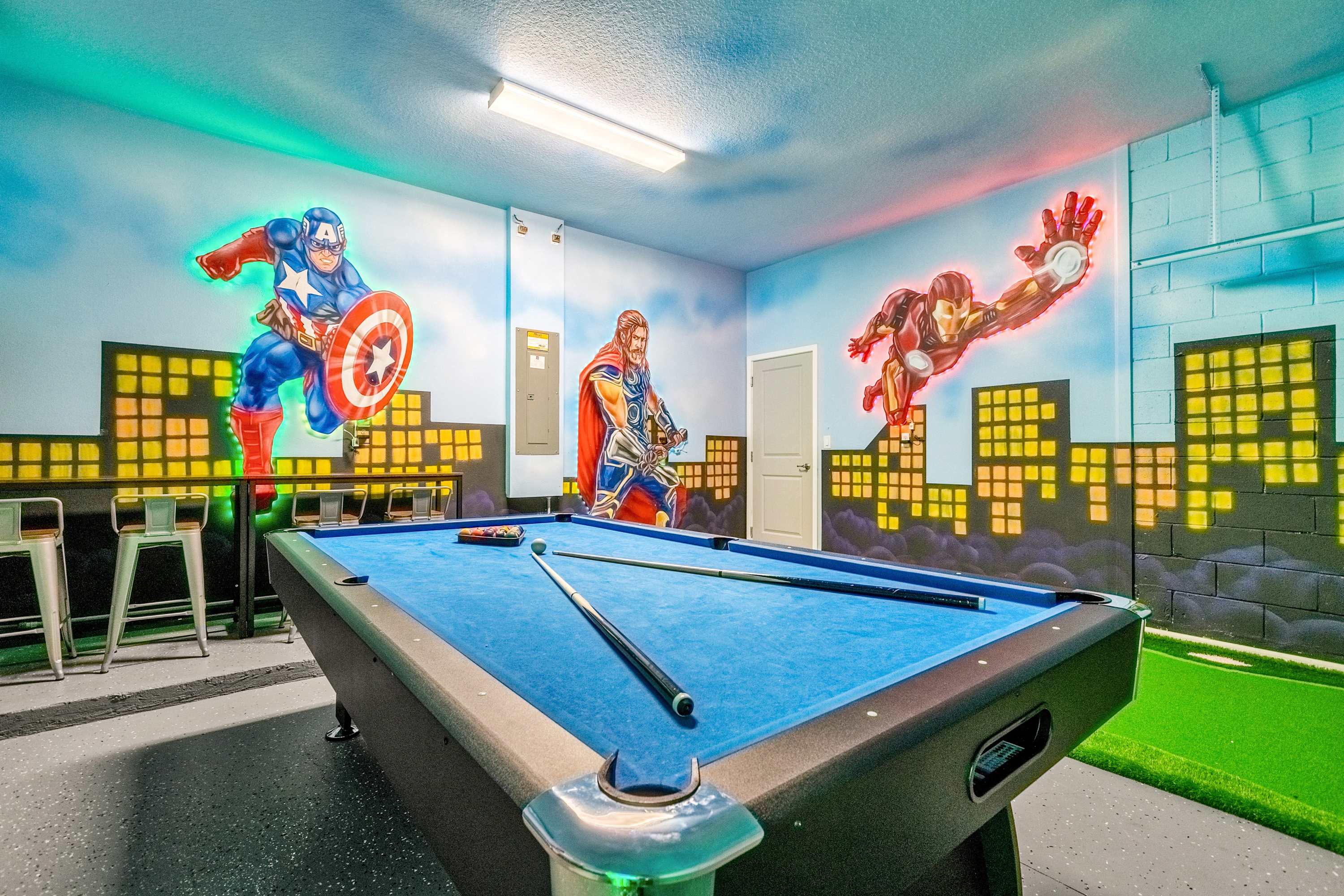 A vibrant game room features a blue-felted pool table in the foreground, with walls adorned with colorful superhero murals. A putting green is visible in the background, adding to the recreational activities.