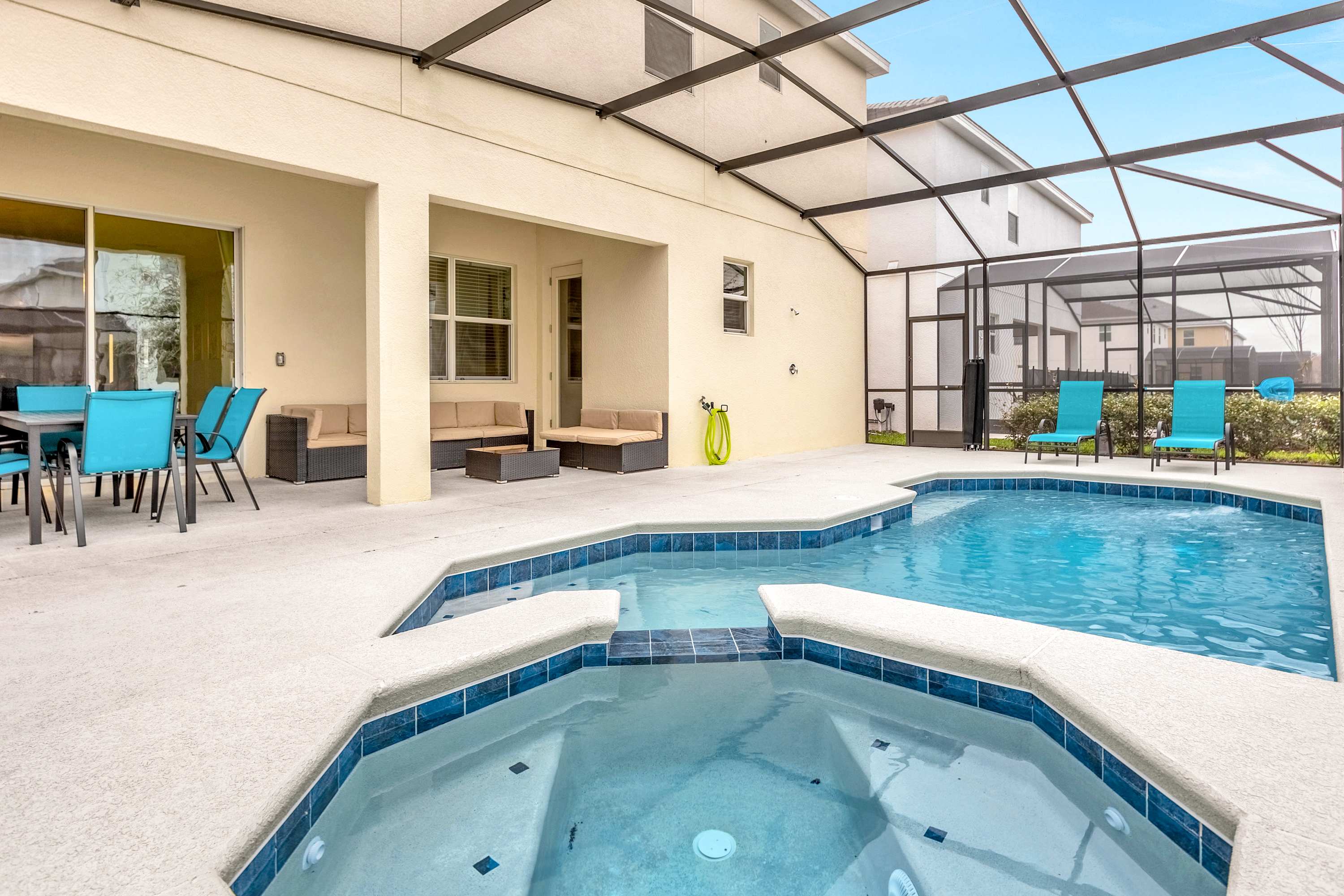 An outdoor screened-in pool area features a large swimming pool with an integrated hot tub, surrounded by a light-colored concrete deck. A covered patio area with outdoor seating and dining furniture is visible on the left.