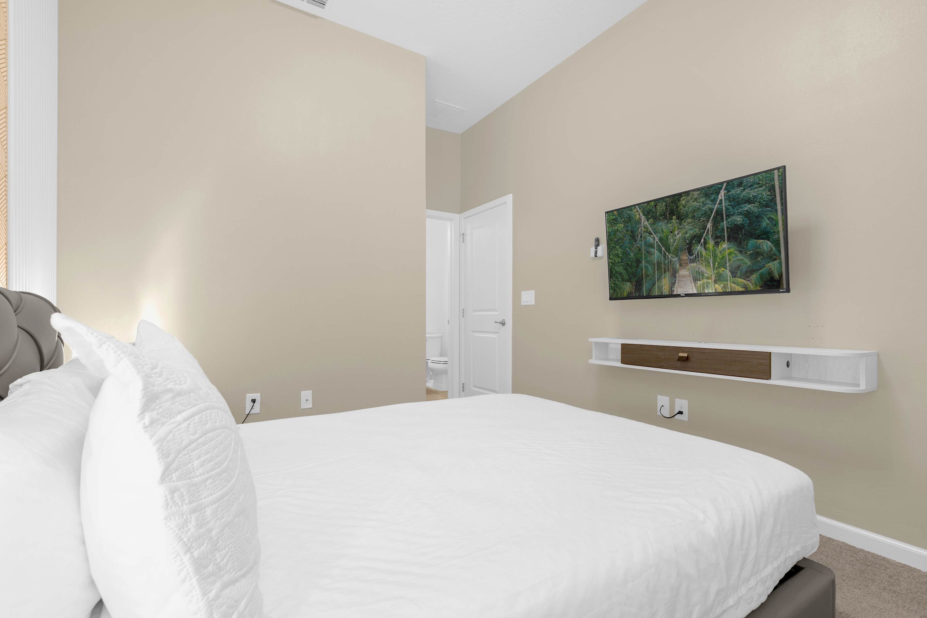 Cozy bedroom featuring a bed with white bedding, a wall-mounted television, and a floating shelf below, with a doorway leading to an adjacent bathroom visible in the background.