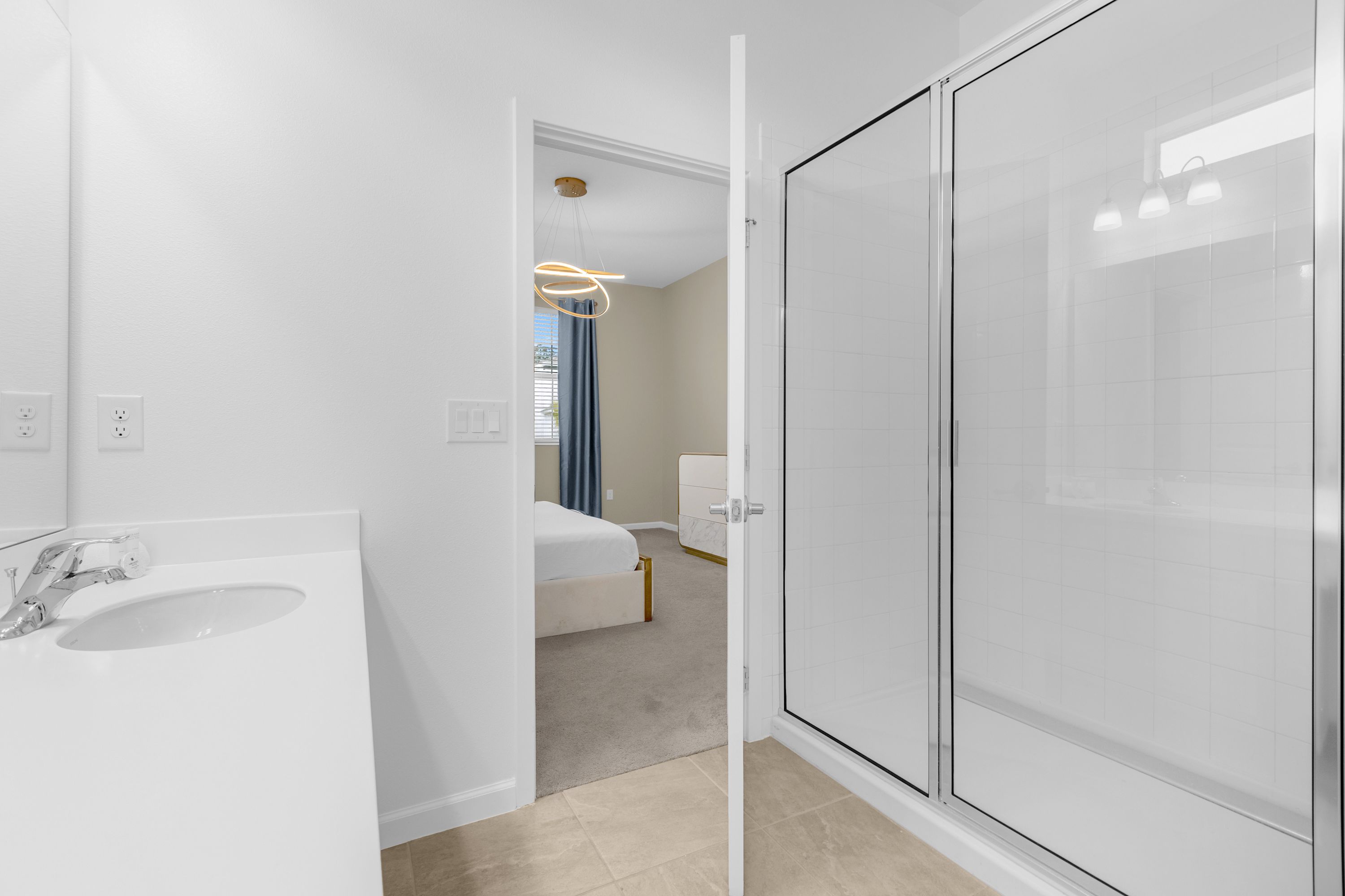 Modern white bathroom featuring a glass-enclosed shower with white subway tiles, a vanity with a single sink, and a glimpse into a bedroom with a round bed and gold chandelier.