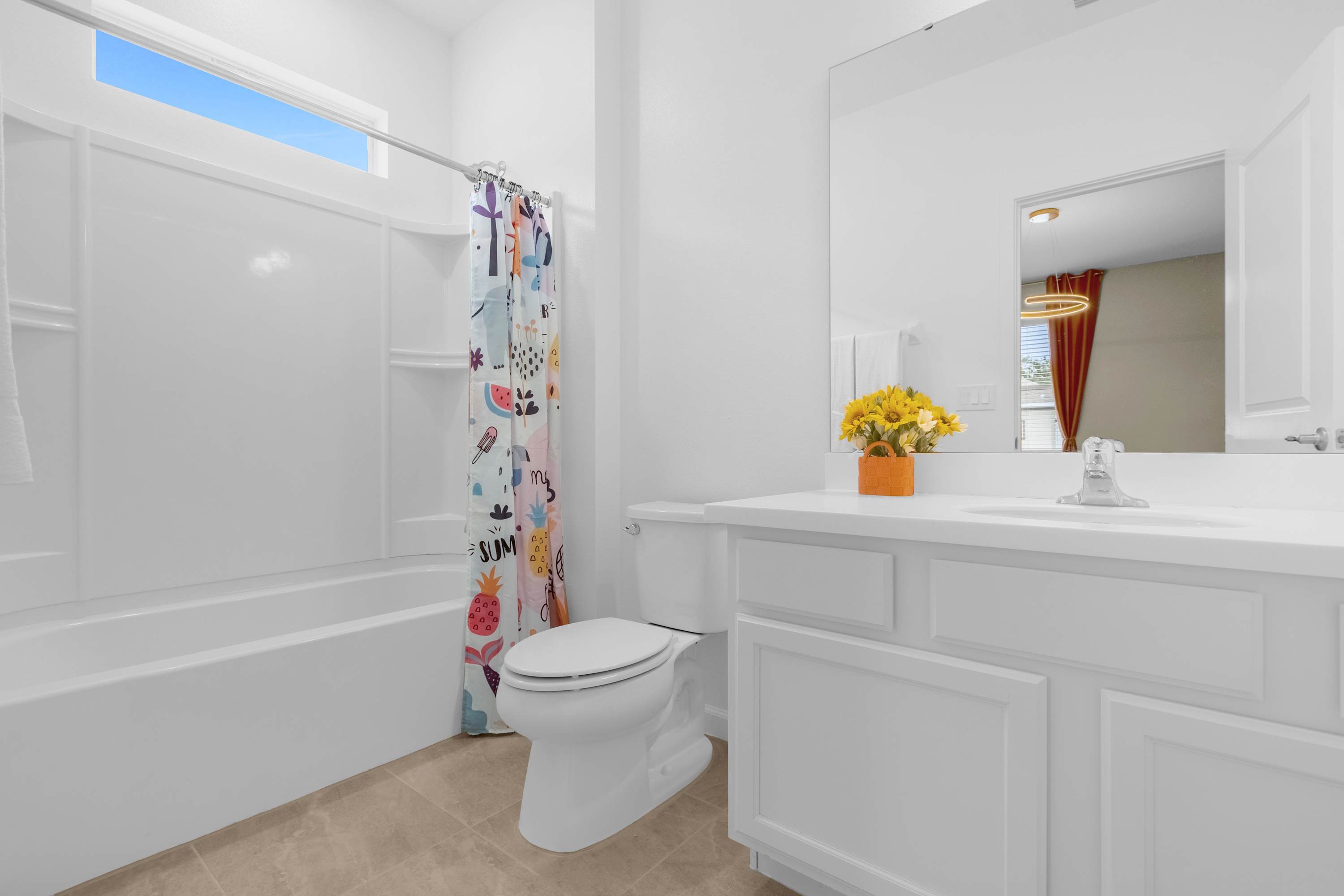 A bright, white bathroom featuring a single vanity with a white countertop and cabinets, a toilet, and a white tub/shower combo with a playful, fruit-patterned shower curtain. A yellow floral arrangement adds a pop of color to the vanity.