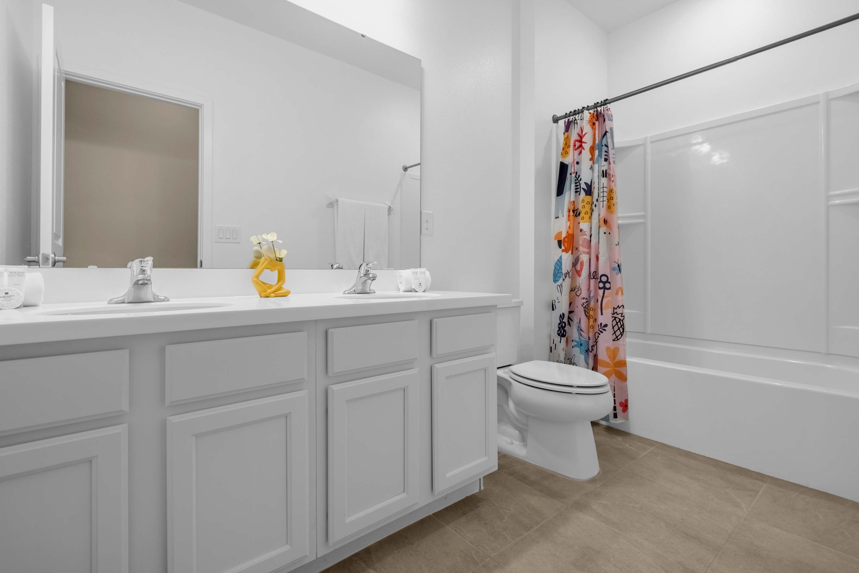 A bright white bathroom featuring a double vanity with two sinks, a large mirror, and a toilet next to a bathtub with a colorful floral shower curtain.