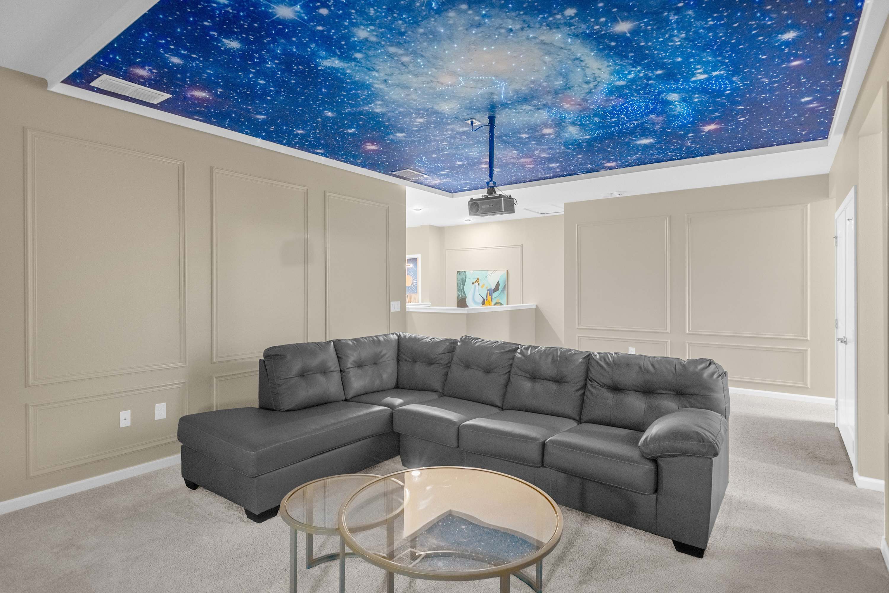 A dedicated media room featuring a large grey sectional sofa and a projector mounted to the ceiling. The ceiling is painted with a vibrant galaxy mural, creating an immersive viewing experience, and the room has carpeted flooring.