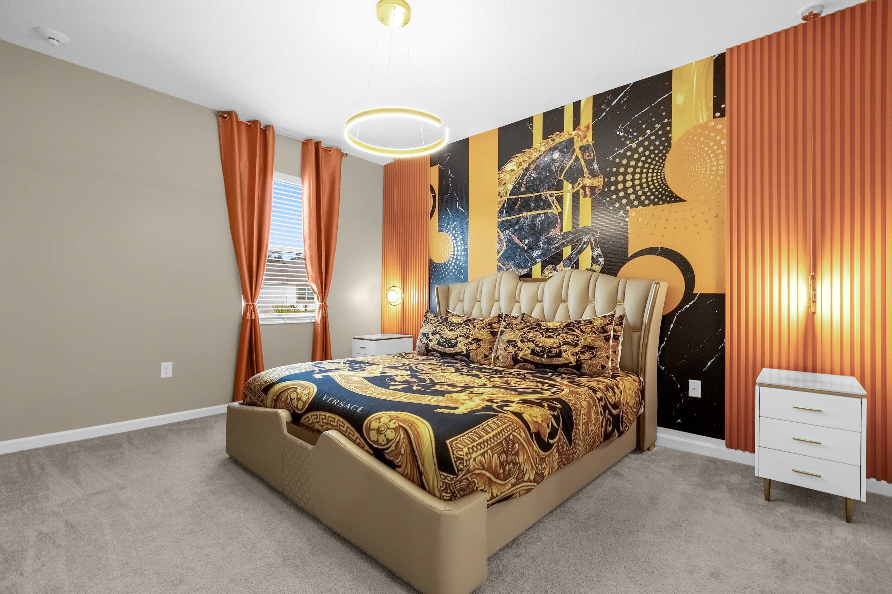 A luxurious bedroom featuring a king-sized bed with a gold upholstered headboard and black and gold patterned bedding. The accent wall behind the bed showcases a bold black and gold mural with a horse motif, flanked by orange fluted panels and hanging lights.
