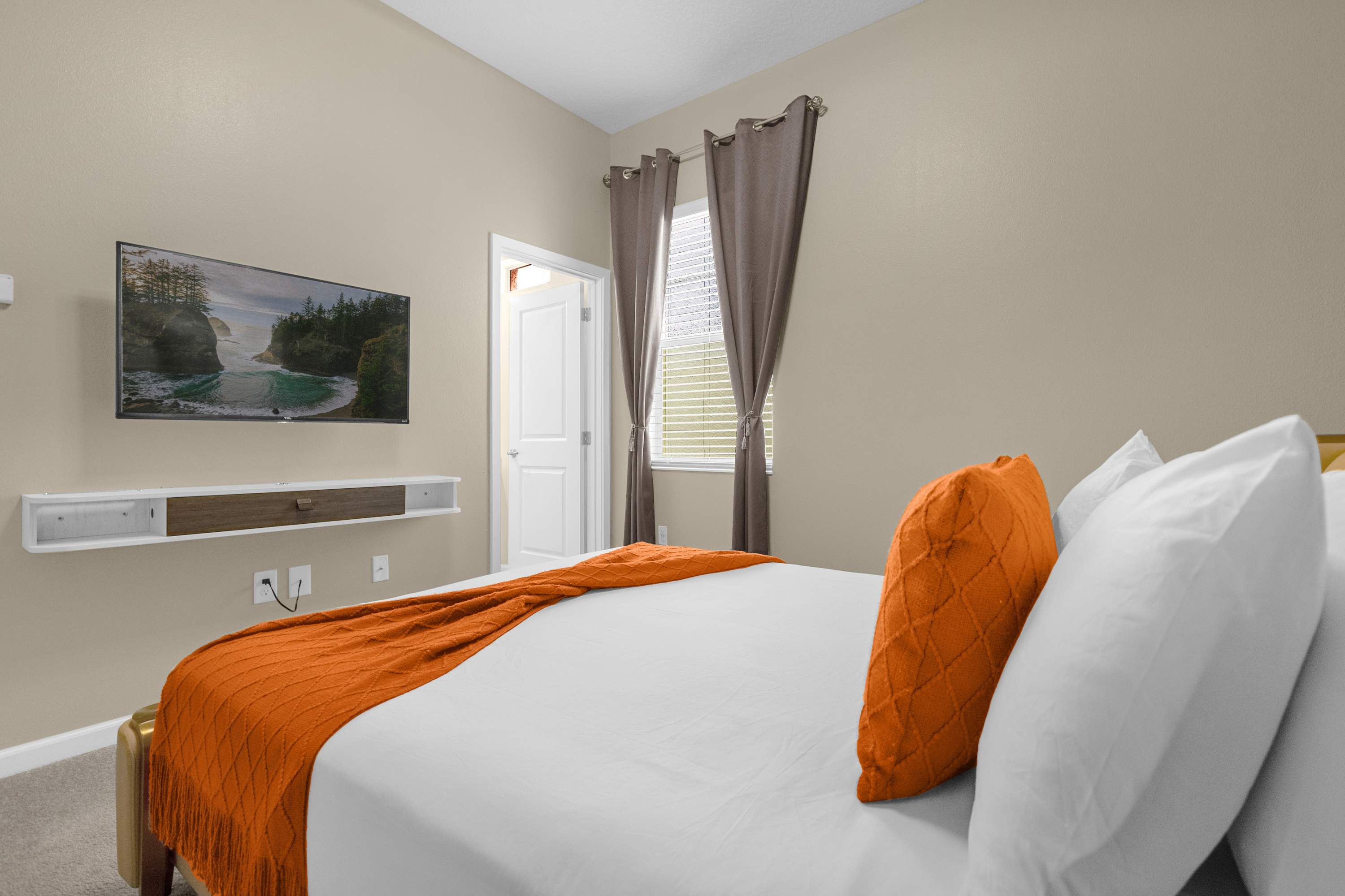 View of the stylish bedroom showing the queen bed with orange accents, a wall-mounted TV above a floating shelf, and a window with dark grey curtains, with light beige walls.