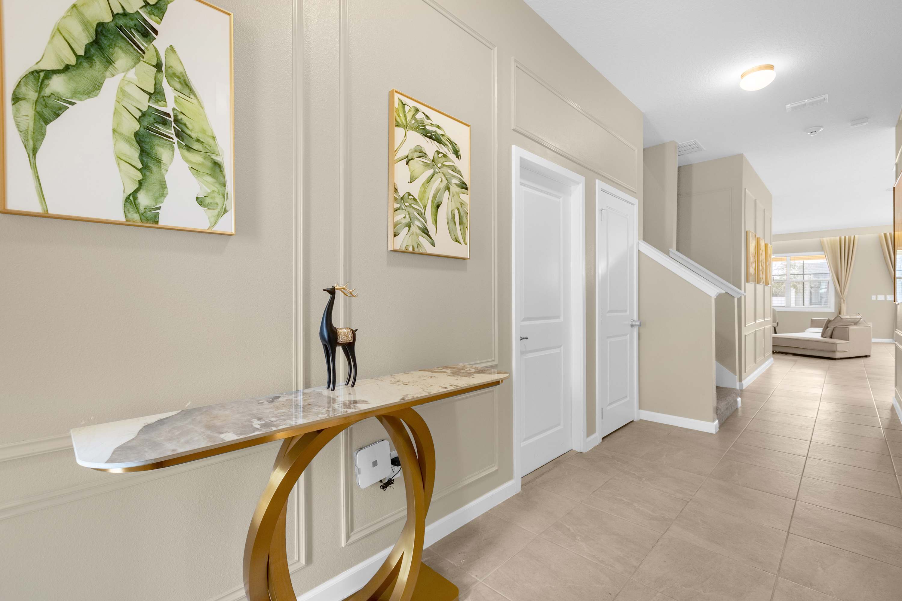 Inviting hallway with light beige walls featuring decorative wainscoting, a modern console table with a marble top and gold base, and tropical leaf artwork, leading towards a staircase.