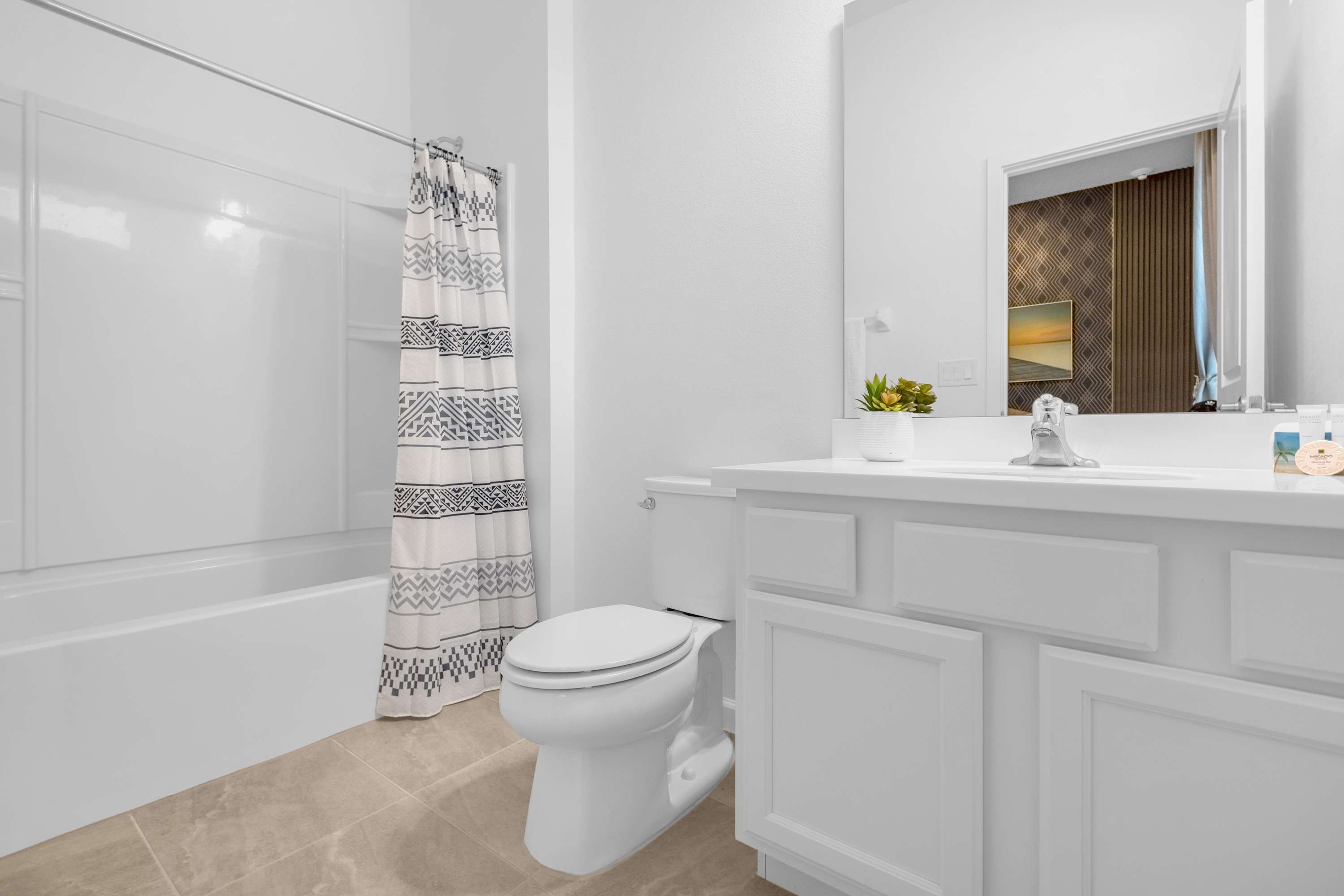A bright, modern bathroom featuring a white vanity with a single sink, a toilet, and a white tub/shower combo with a black and white patterned shower curtain. Light beige tiled floors complete the clean aesthetic.