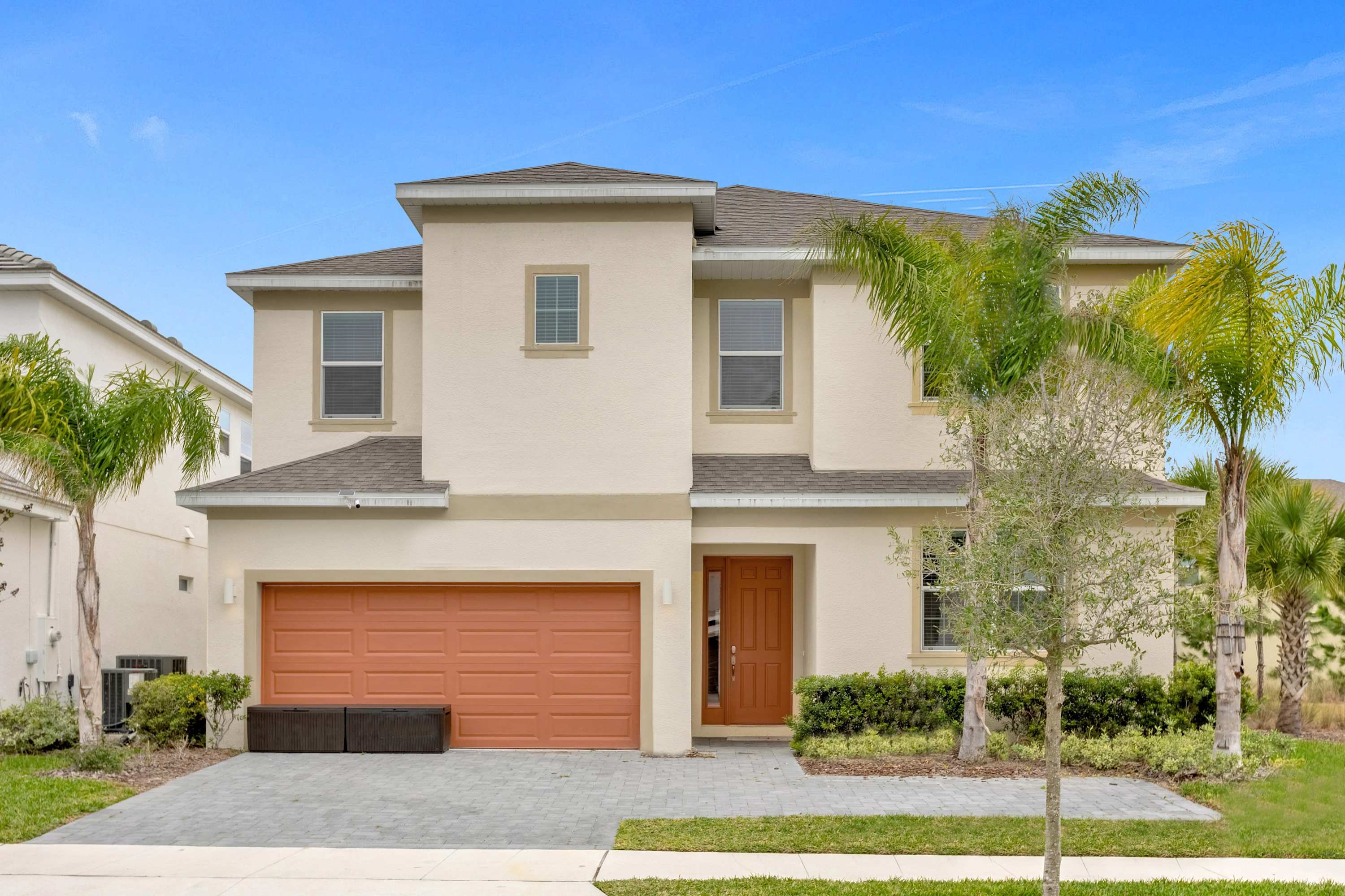 Modern two-story house with a light beige stucco exterior, an orange garage door, a matching front door, and a paved driveway, flanked by palm trees and landscaping under a clear blue sky.