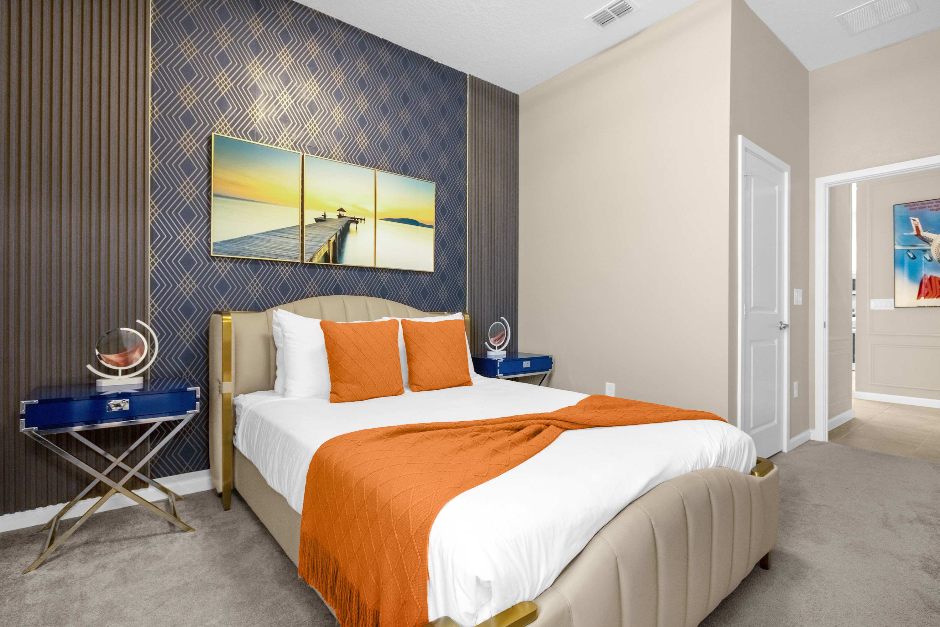 Stylish bedroom with a queen-sized bed, a dark blue geometric accent wall with gold trim, and three framed pictures of a pier at sunset, flanked by modern nightstands.